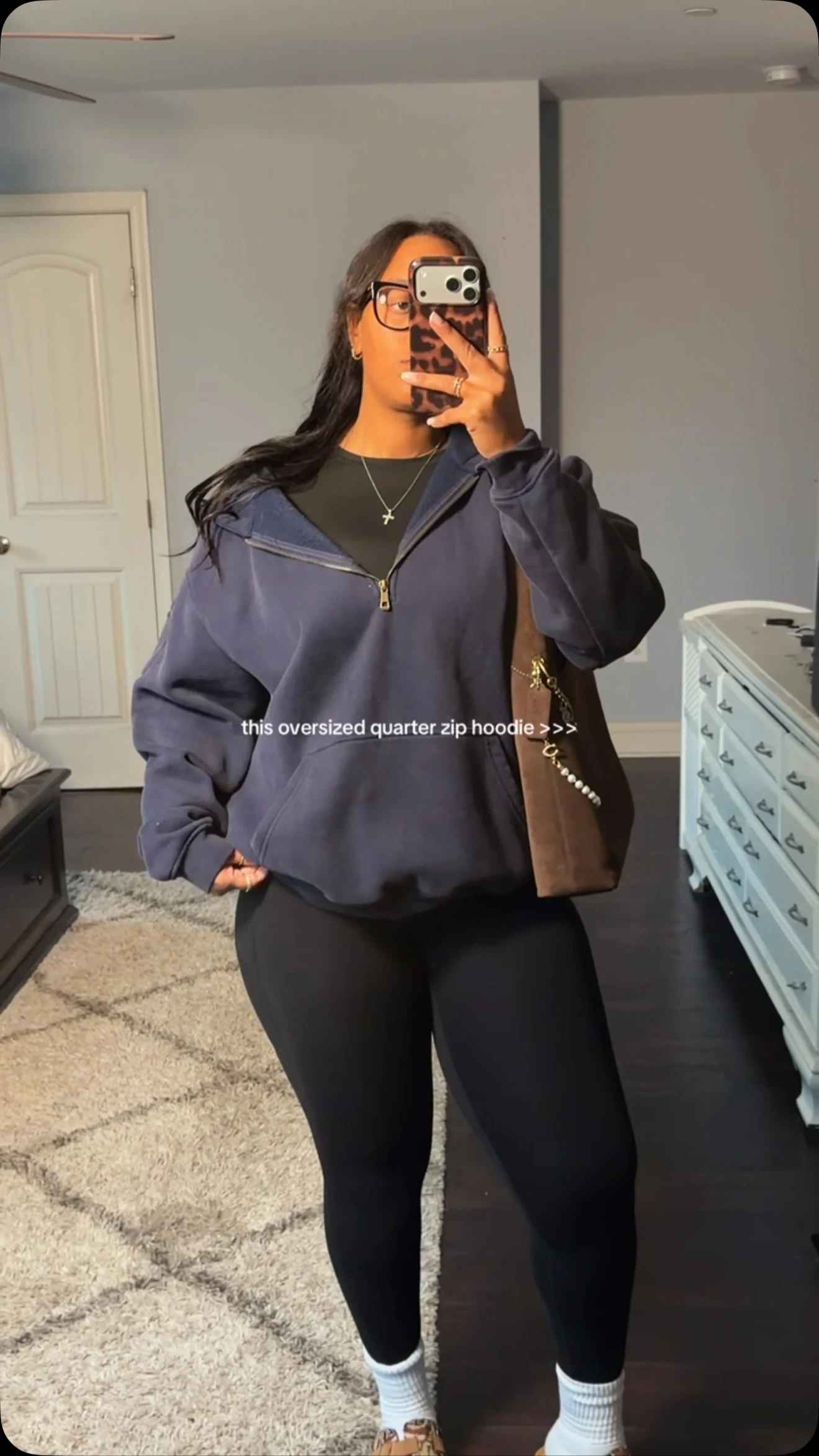 Woman in an oversized navy quarter-zip hoodie, black leggings, leopard slip-ons, and a brown shoulder bag.
