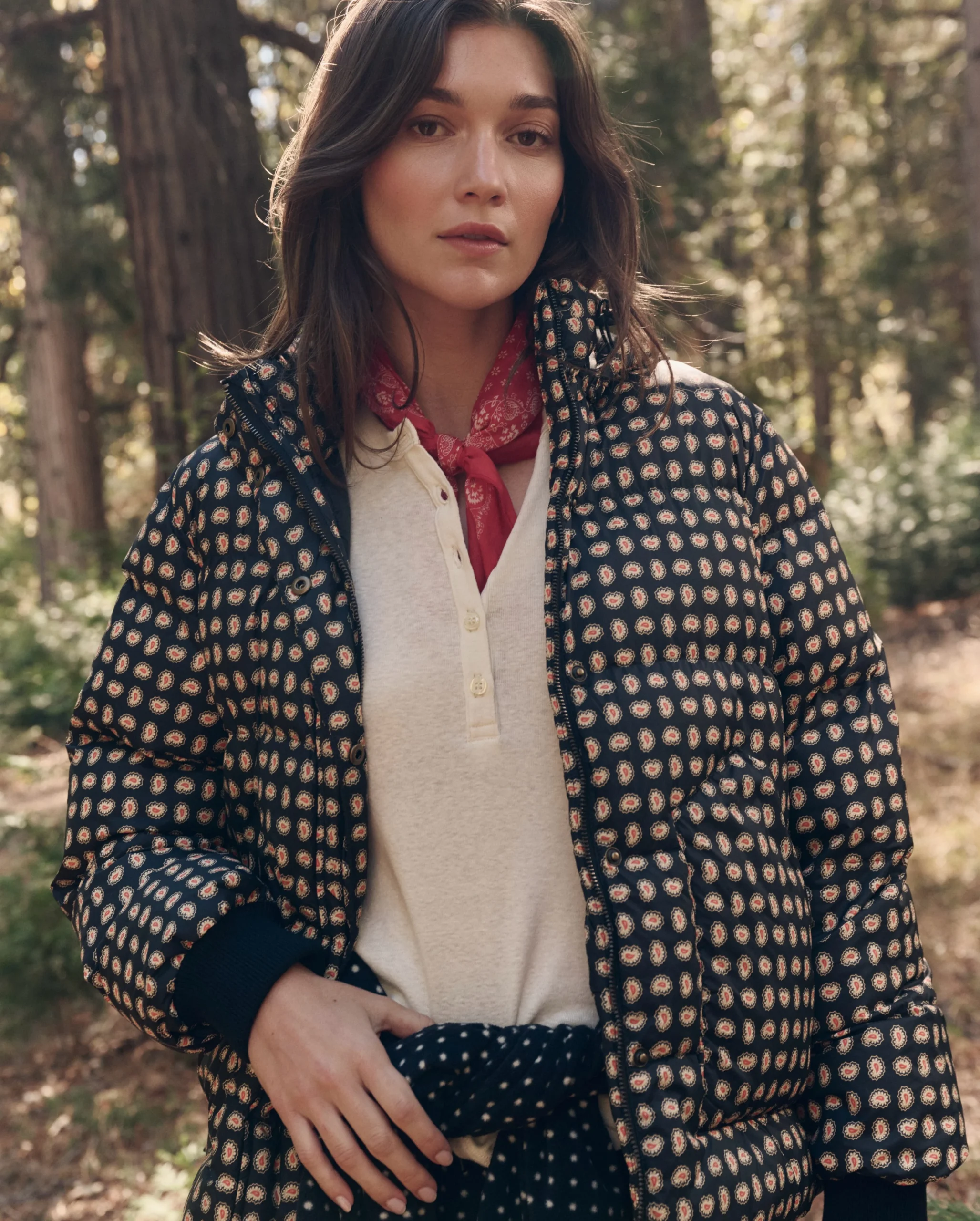 Person wearing a patterned quilted jacket belted at the waist with a red scarf, outdoors among trees.
