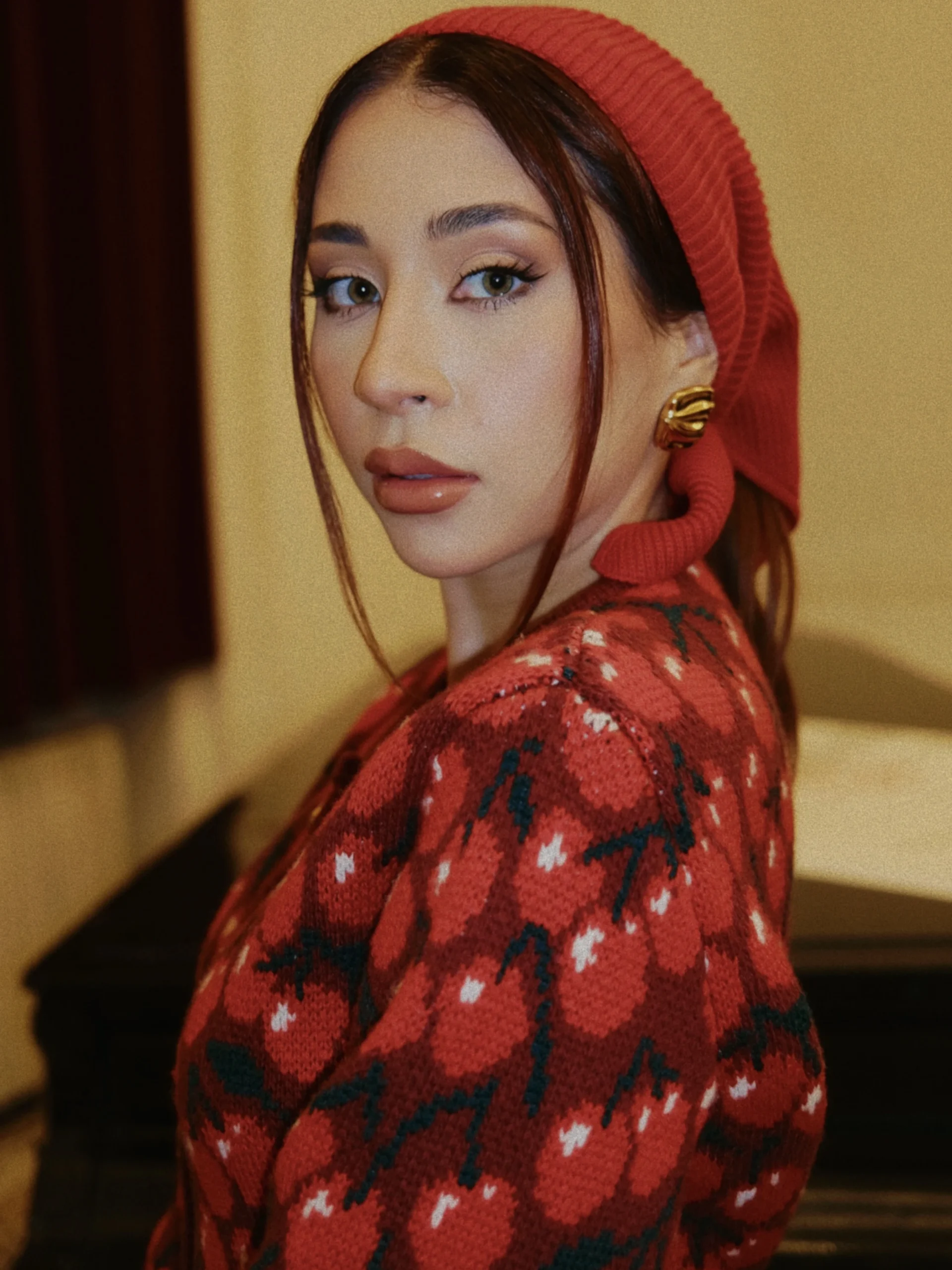Woman wearing a red ribbed beanie and a red cherry-pattern sweater with a gold sculptural earring.