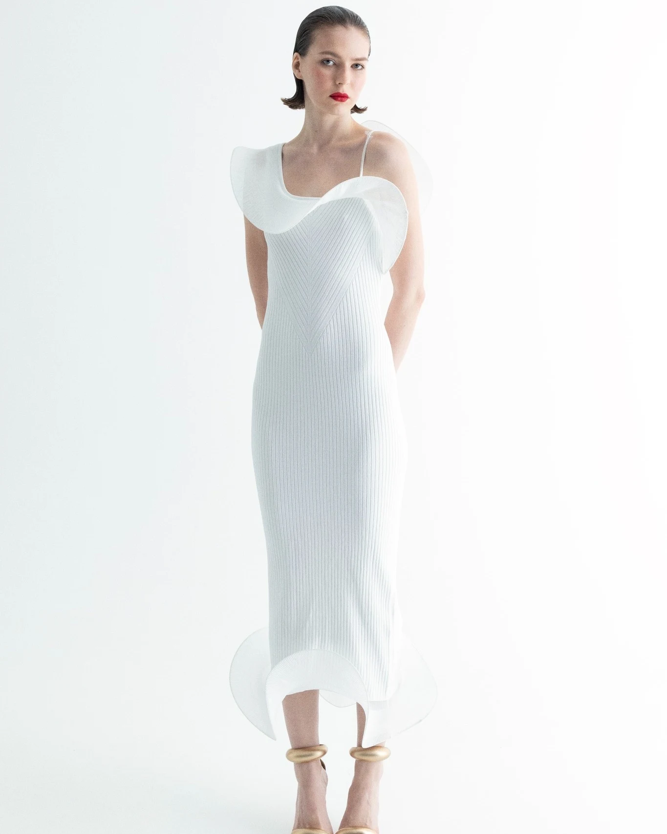 White ribbed midi dress with sculptural curved panels and gold ring ankle accessories on a model.