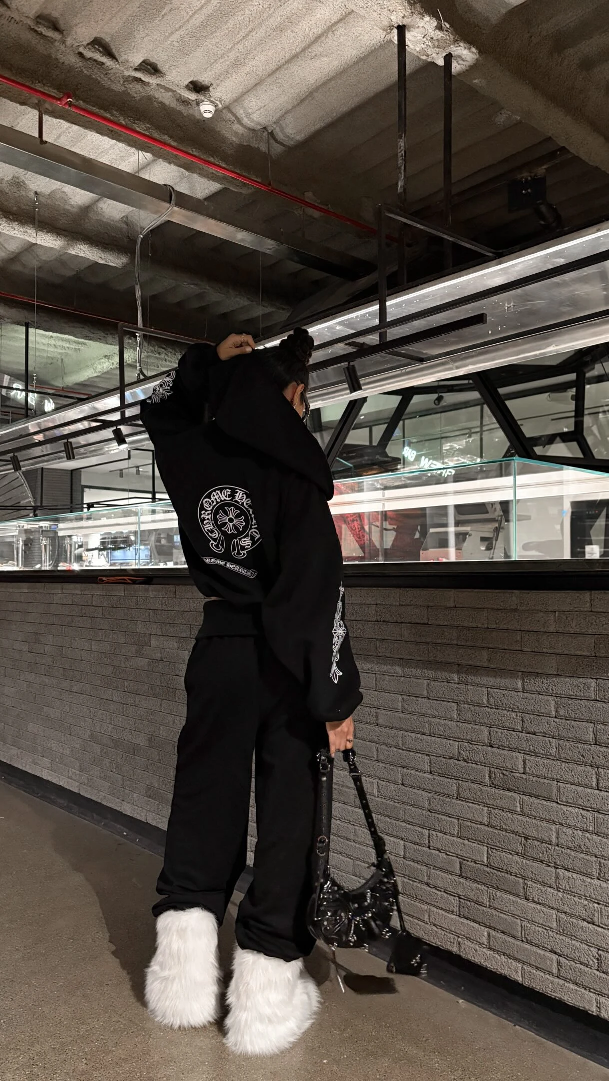 Person in a black graphic hoodie, wide-leg pants, and oversized white furry shoes holding a black bag.