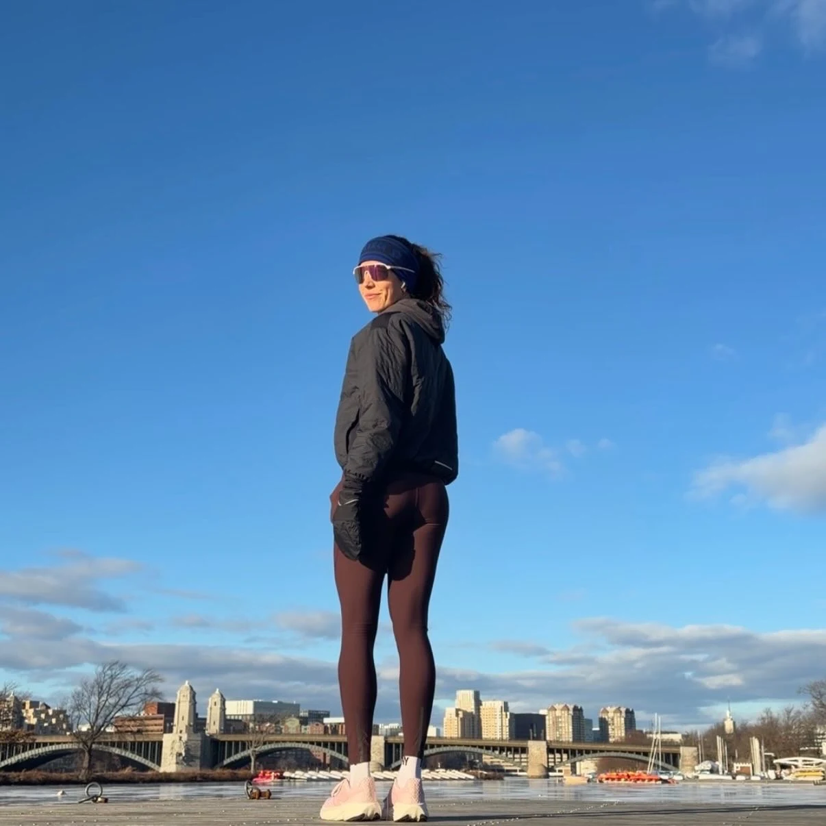 Black cropped hooded jacket with dark leggings, light shoes, headband, and pink sunglasses.