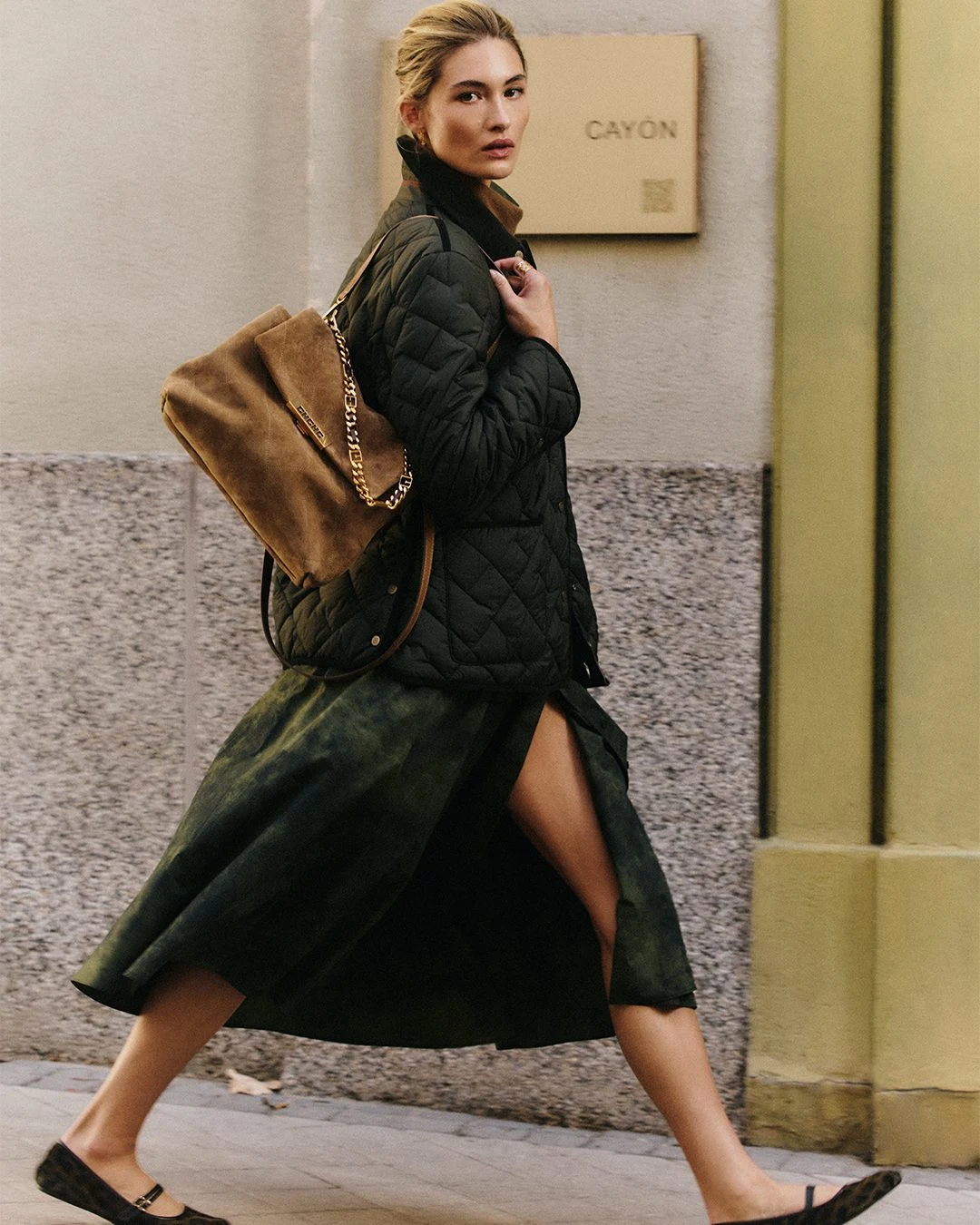 Person in black quilted jacket, green flared skirt, and brown shoulder bag walking near a stone wall.