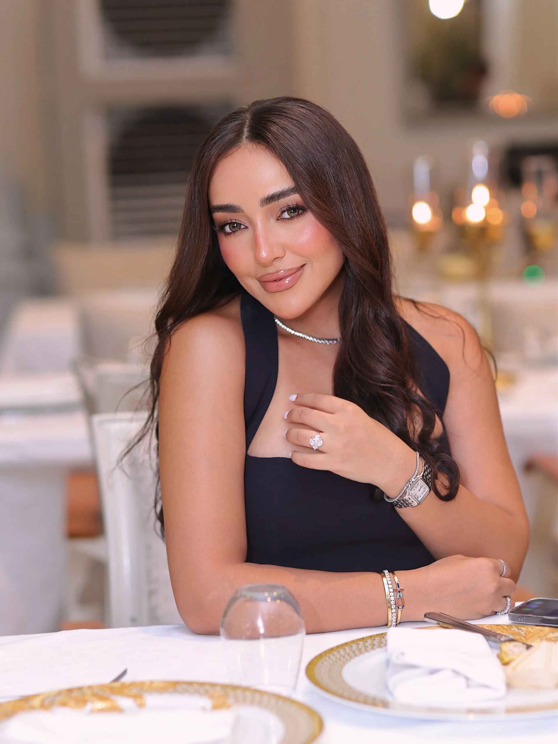 Woman in a fitted black sleeveless dress with a deep neckline seated at a gold-rimmed dinner table.