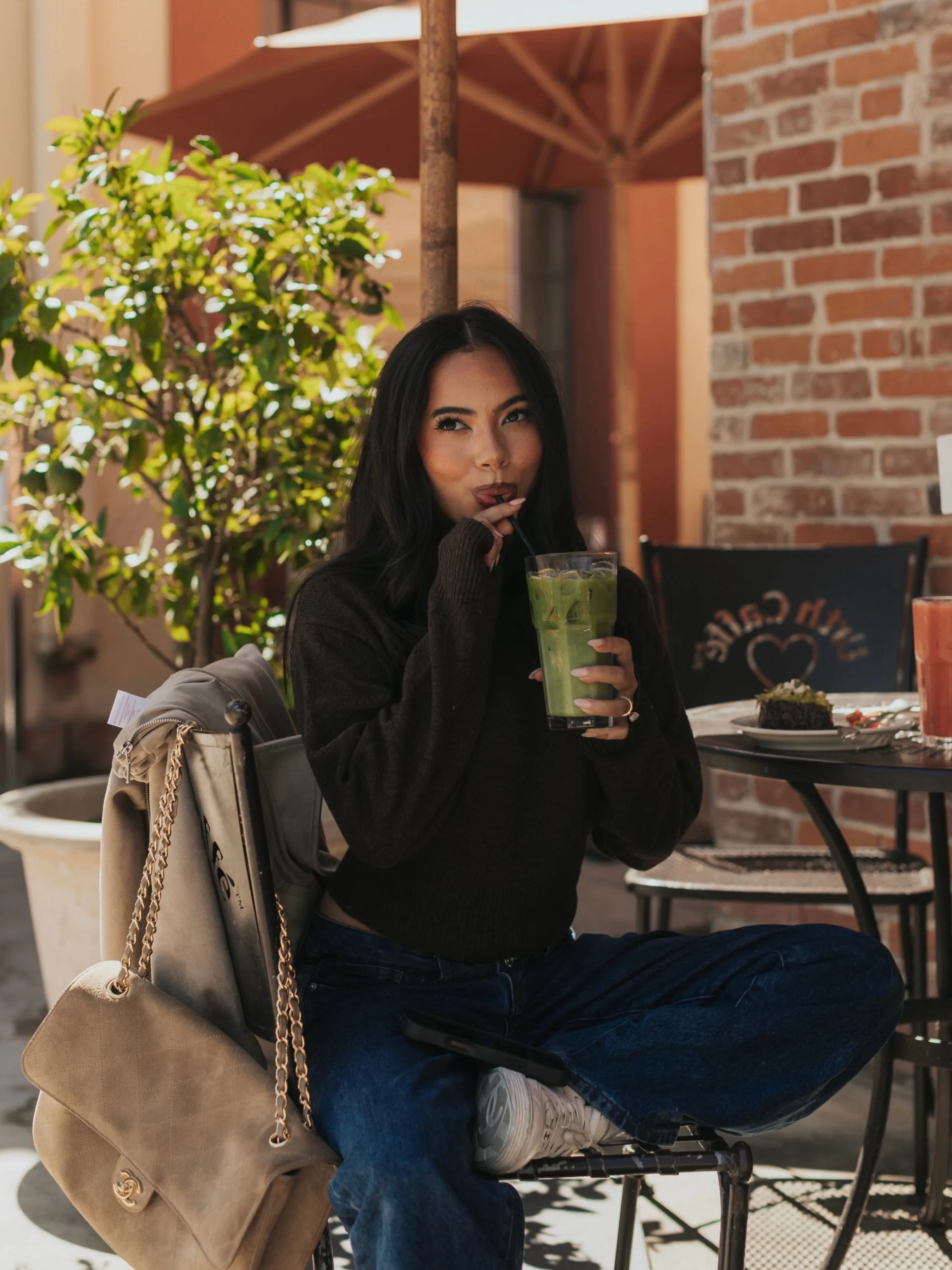 Person seated cross-legged in black sweater, blue jeans, white sneakers, and a beige chain-strap bag.