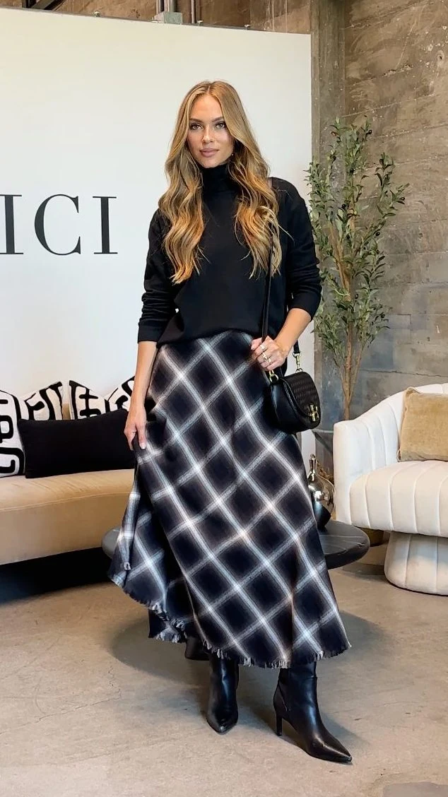 A fitted black long-sleeve turtleneck is tucked into a flowing, ankle-length skirt with a large black, gray, and white plaid pattern and a slightly frayed hem. Black pointed-toe ankle boots are visible beneath the skirt, and a small black shoulder bag hangs at the wearer’s side.