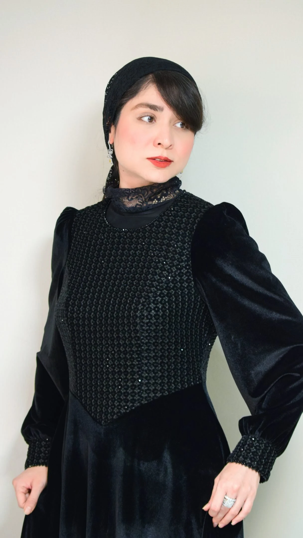 The outfit is a black dress with long puffed sleeves, gathered cuffs, and a fitted bodice featuring a textured, patterned front panel. A sheer black lace layer sits around the high neckline. The skirt continues below the frame, and a black head covering completes the visible look.