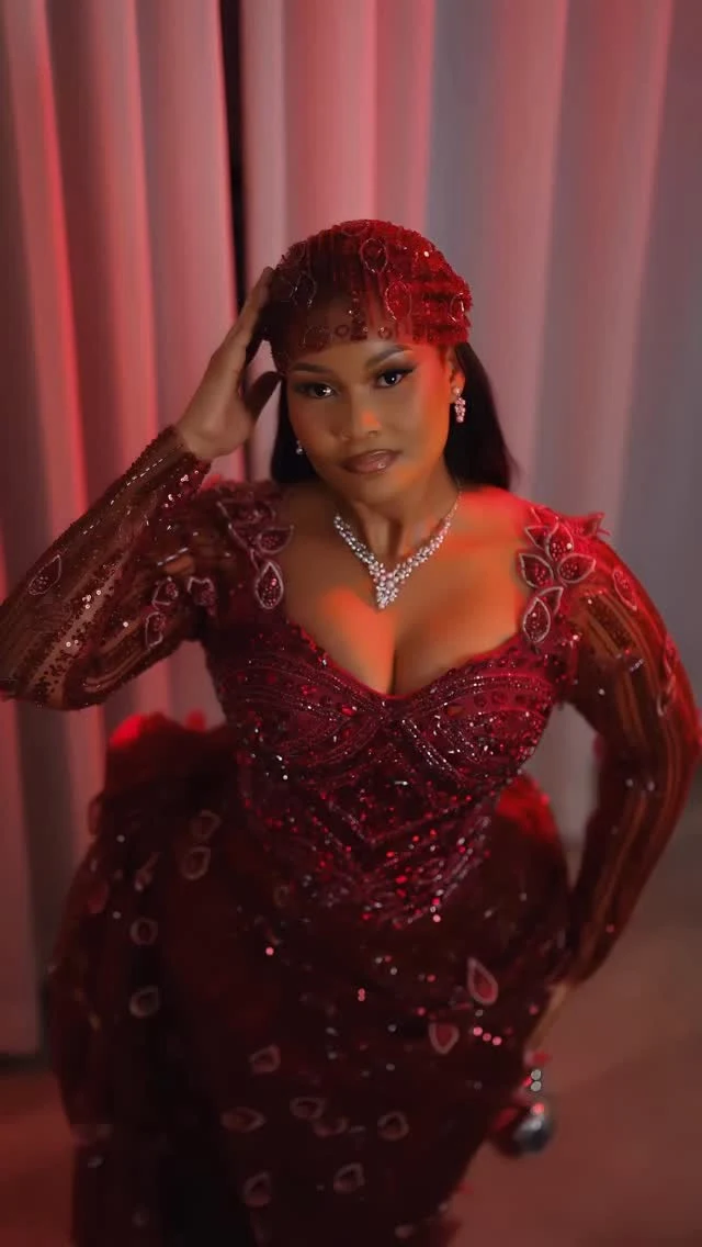 A woman wears a fitted burgundy gown with sheer sleeves, an embellished head covering, and a sparkling necklace.