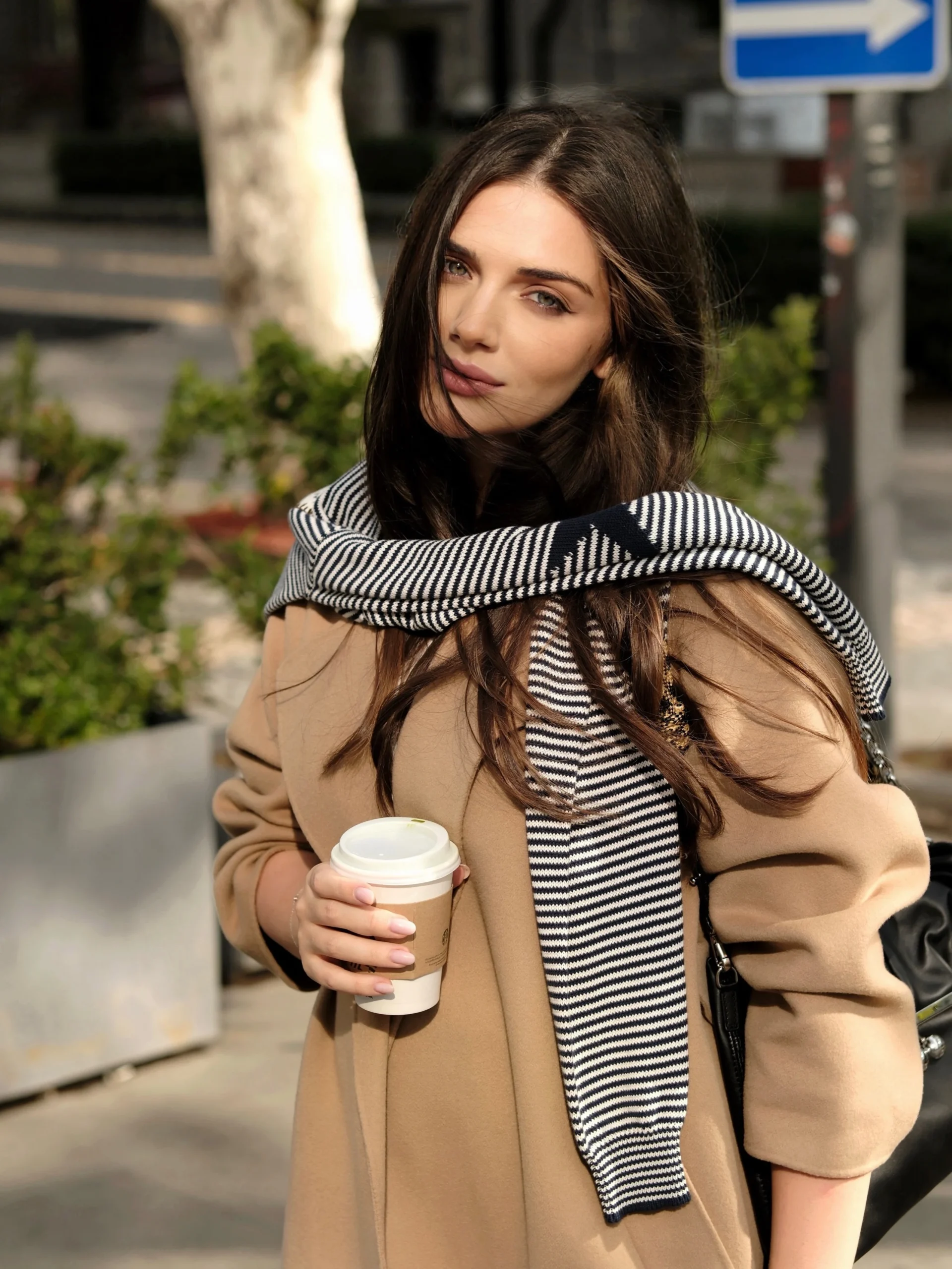 Woman in long camel coat with striped scarf and black shoulder bag holding a white takeaway cup.