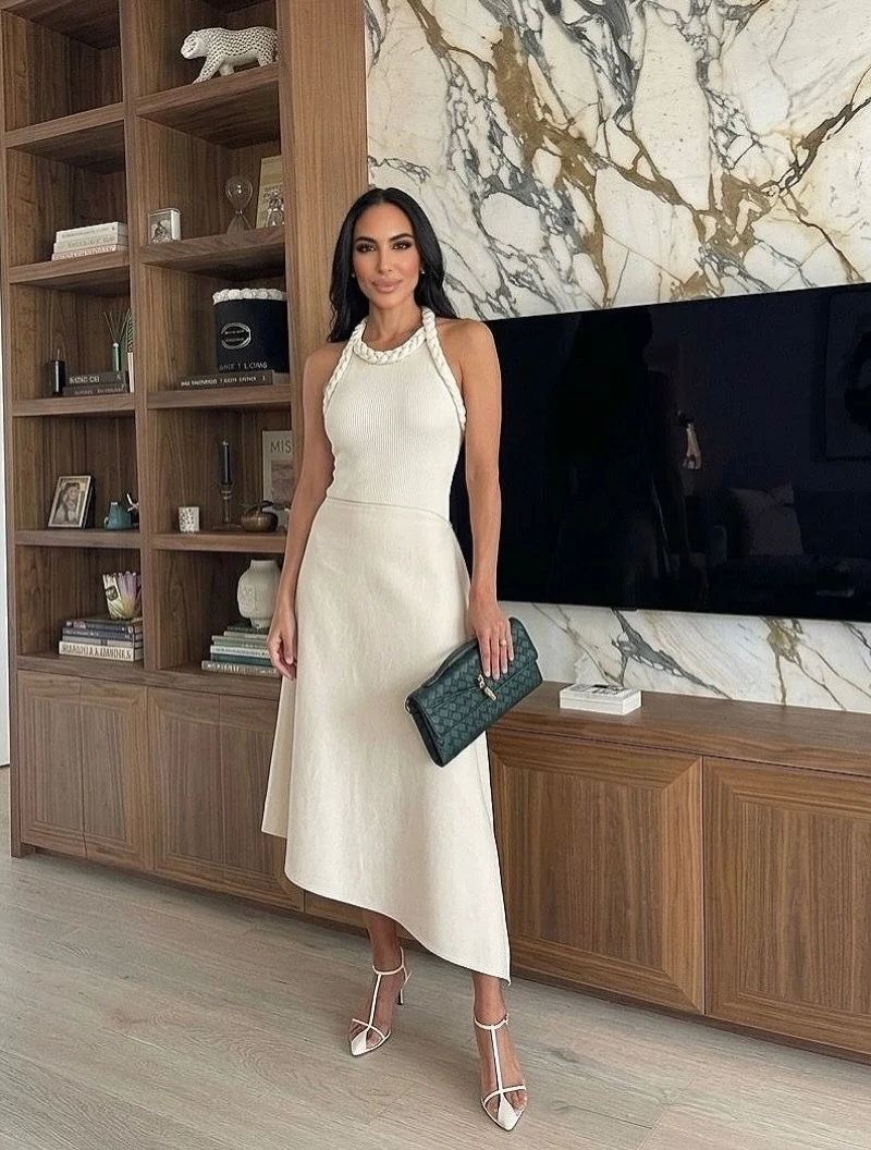 Woman in a cream asymmetrical halter midi dress with white heels and a dark green clutch.