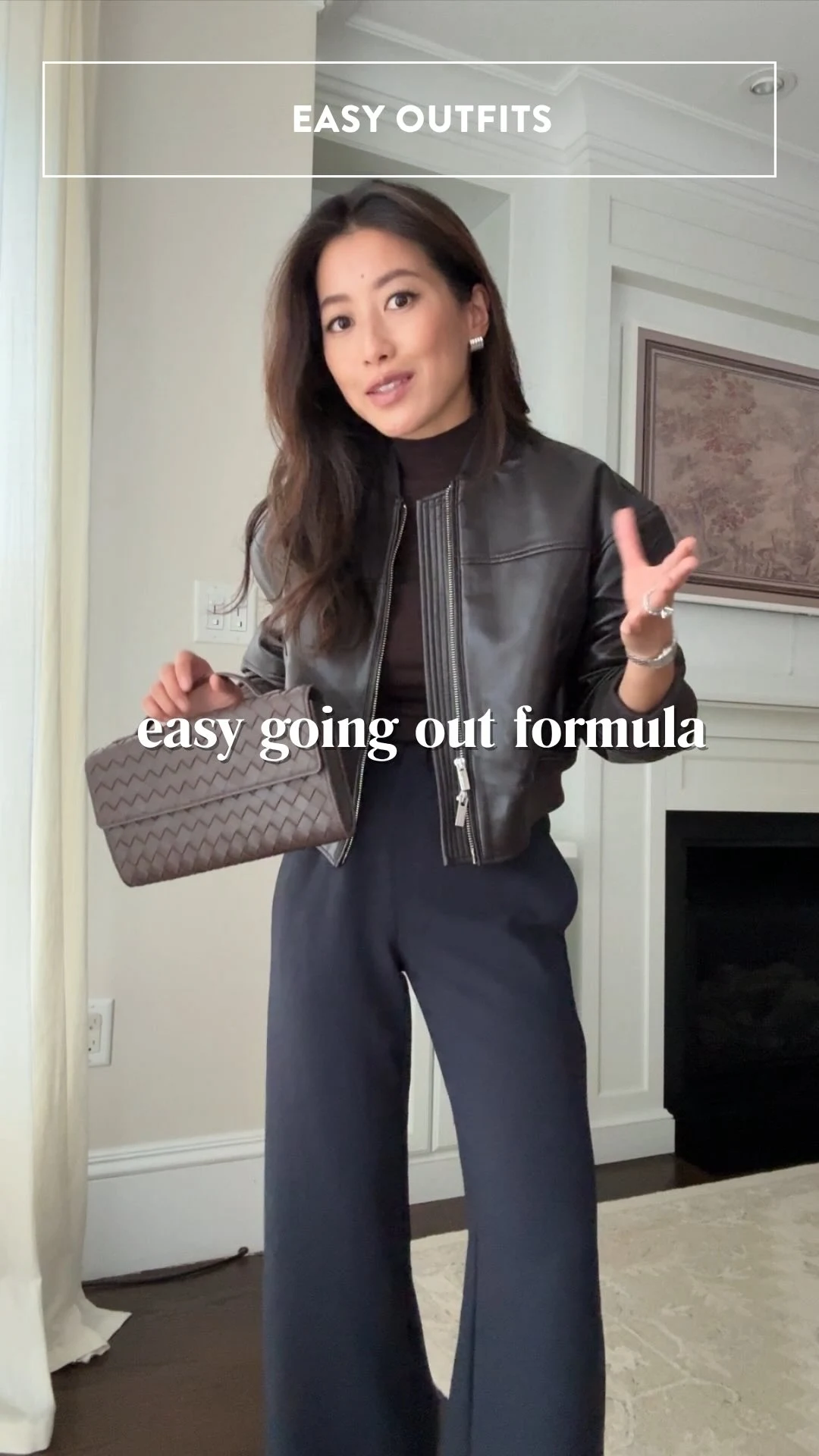 Black cropped zip jacket over dark high-neck top and wide-leg trousers, taupe woven clutch at side.