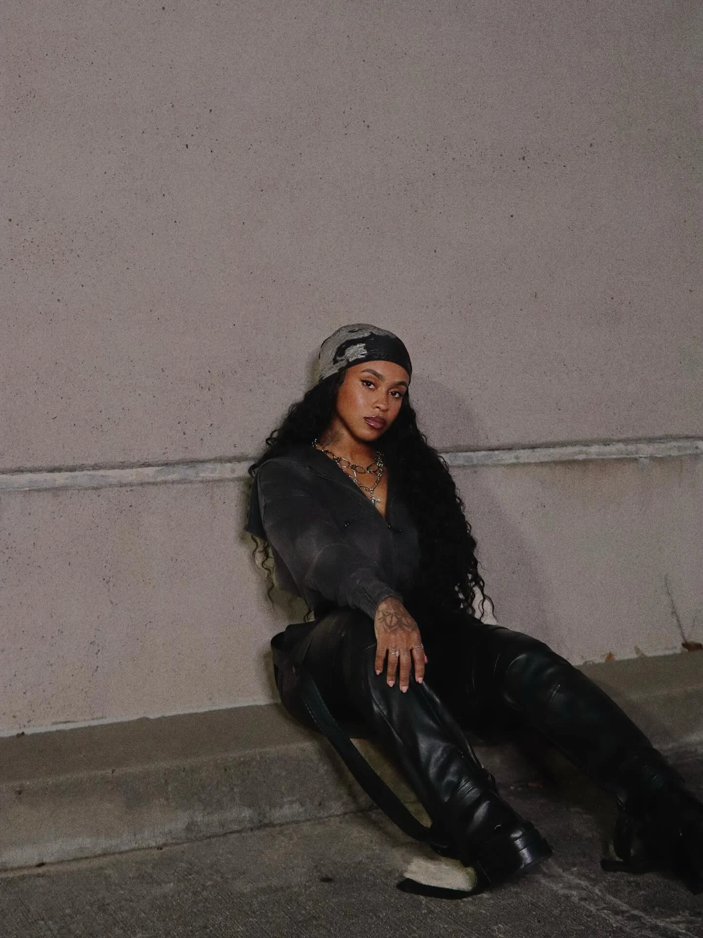 Cropped zip-up black jacket, high-waisted shiny black leather pants with side straps, black platform boots, layered silver chain necklaces, patterned durag