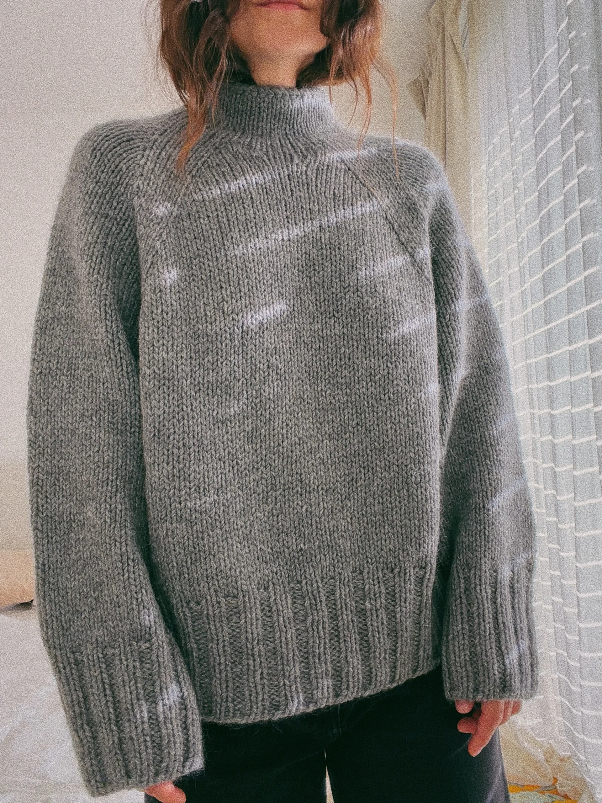 Person wearing a roomy gray turtleneck sweater with ribbed cuffs and dark pants.