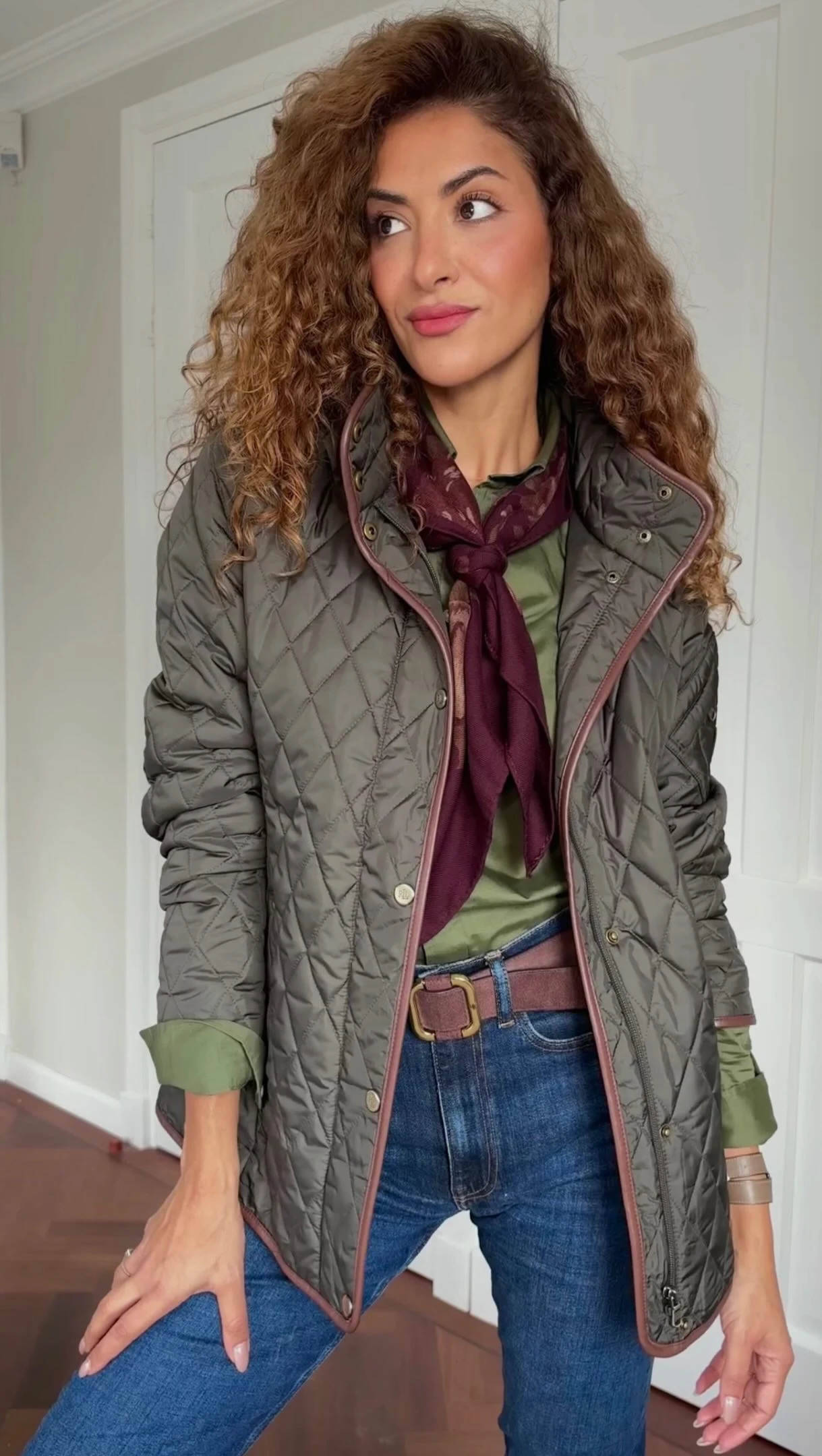Dark blue jeans, olive green button-down shirt, brown belt, tall brown leather riding boots, olive quilted jacket with brown trim, burgundy floral scarf at neck, tan crossbody bag