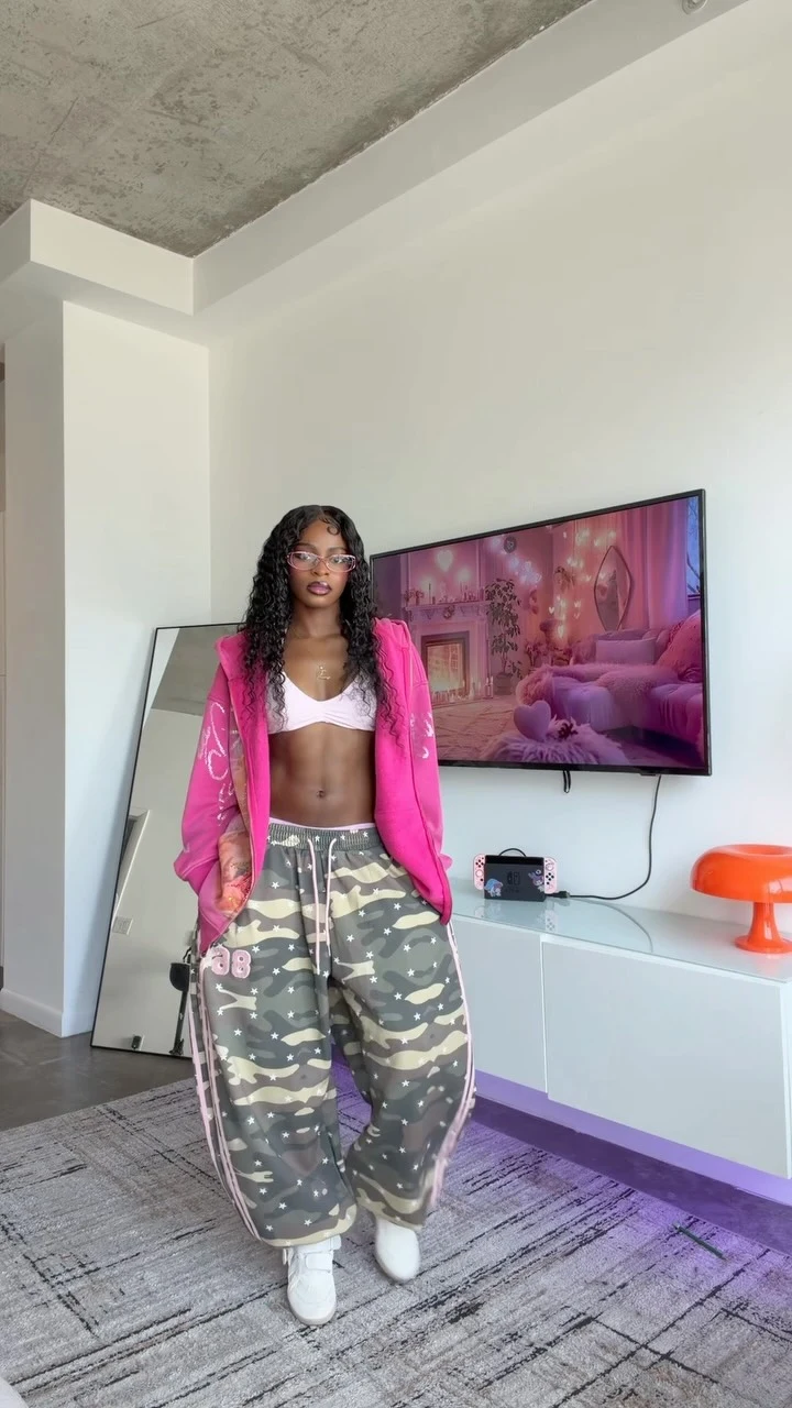 Woman in a bright pink zip-up hoodie, pink bralette, baggy camo cargo pants, and white sneakers.
