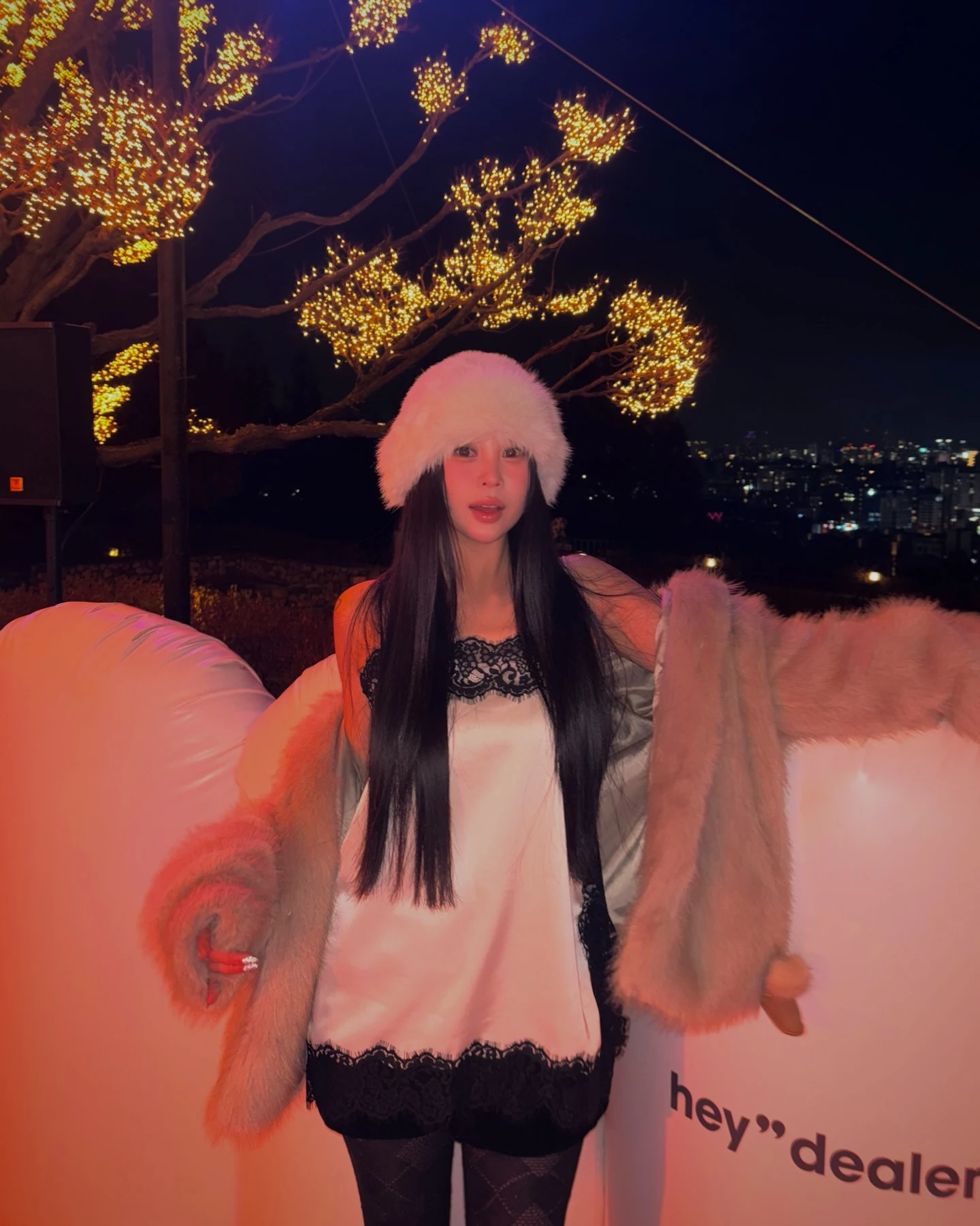 Person in a pale slip dress with black lace trim, blush fur coat, white hat, and sheer tights.
