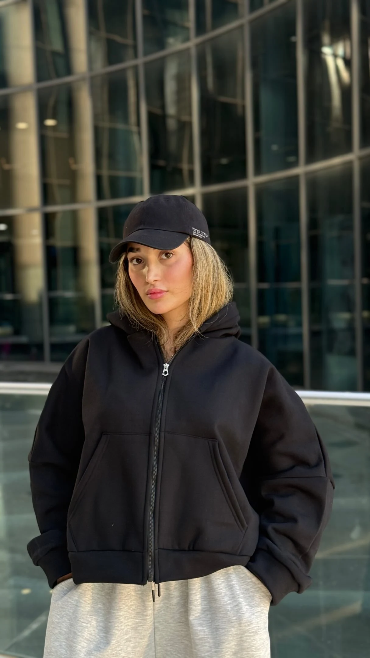 black cropped zip-up hoodie with back text, loose light-grey sweatpants, black visor cap, sneakers