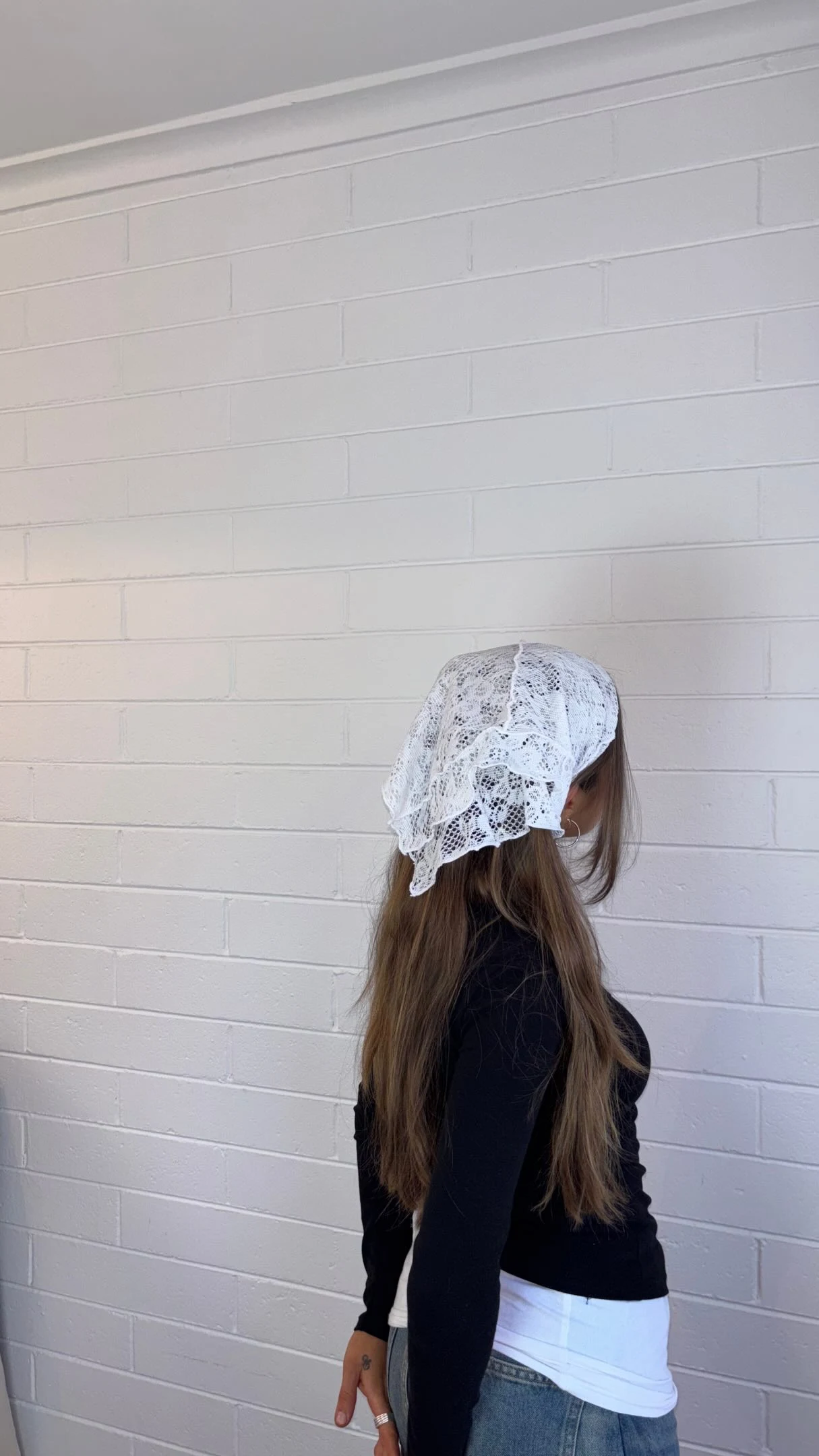 A fitted black long-sleeve top is worn with a white lace headscarf draped over the head. A white layer peeks out below the top, above blue jeans. The person is shown in profile against a pale block wall.