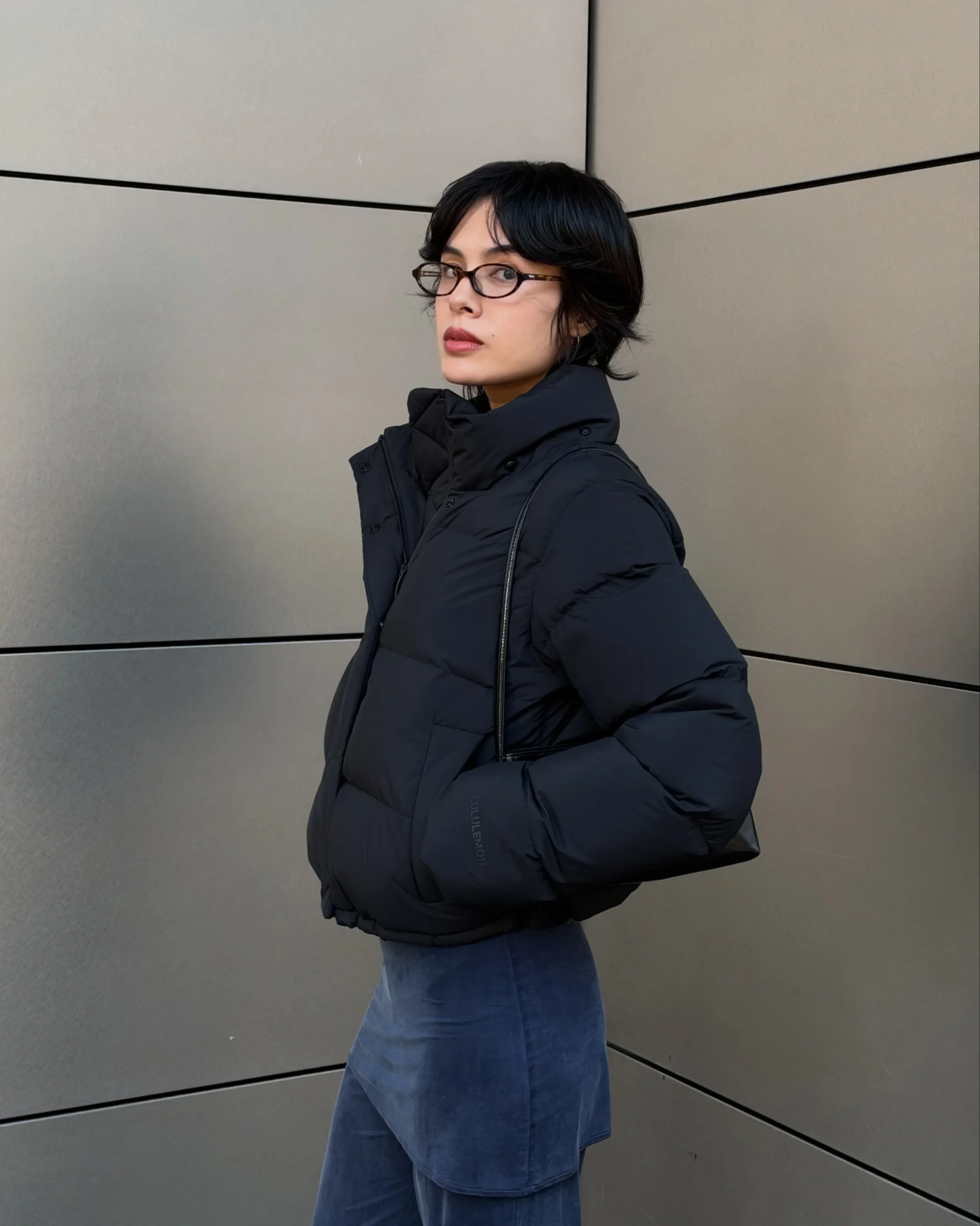 Bulky black puffer jacket with blue trousers and a black shoulder bag tucked under the arm.