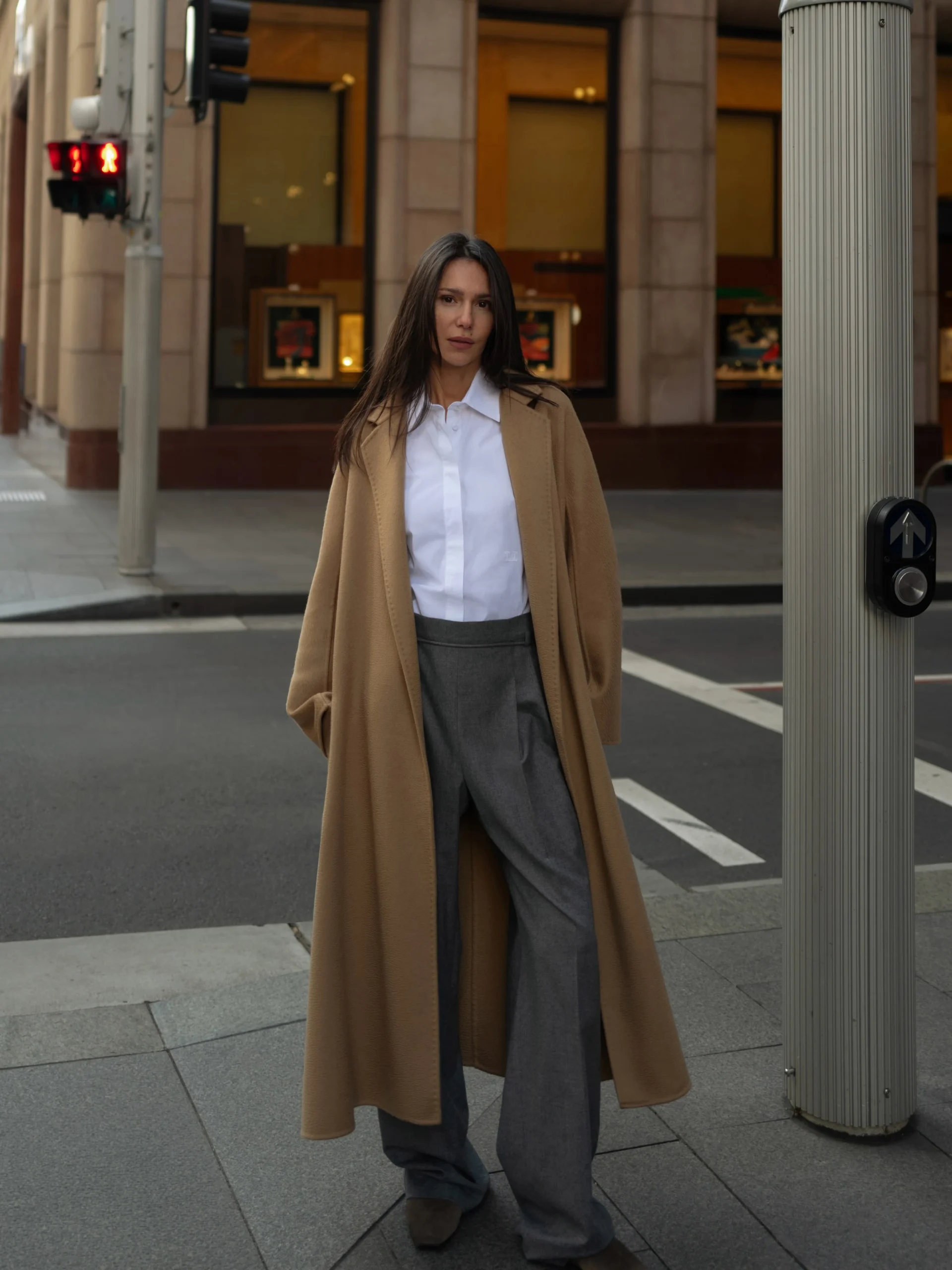 Long camel coat over white shirt and wide-leg gray trousers on a city sidewalk near a crosswalk.