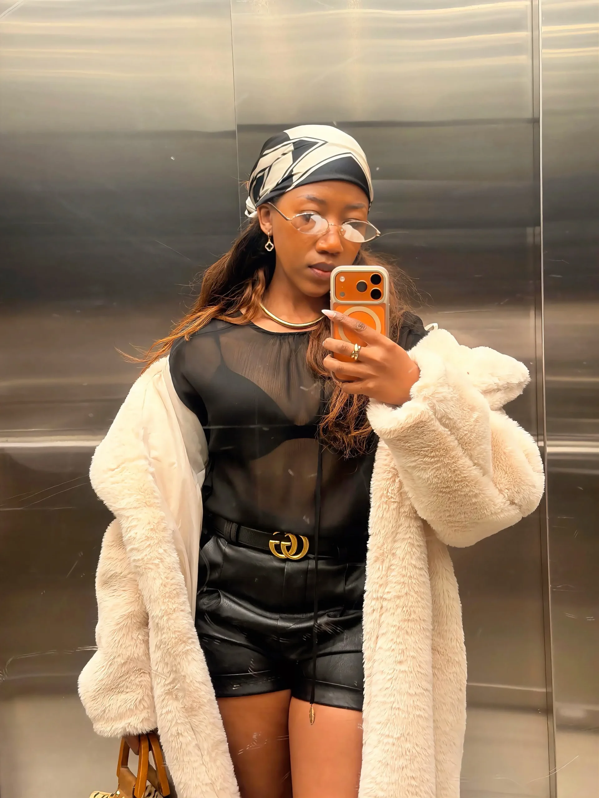 Long cream faux-fur coat, sheer black blouse over black bra, high-waisted black leather shorts, black belt, brown handbag