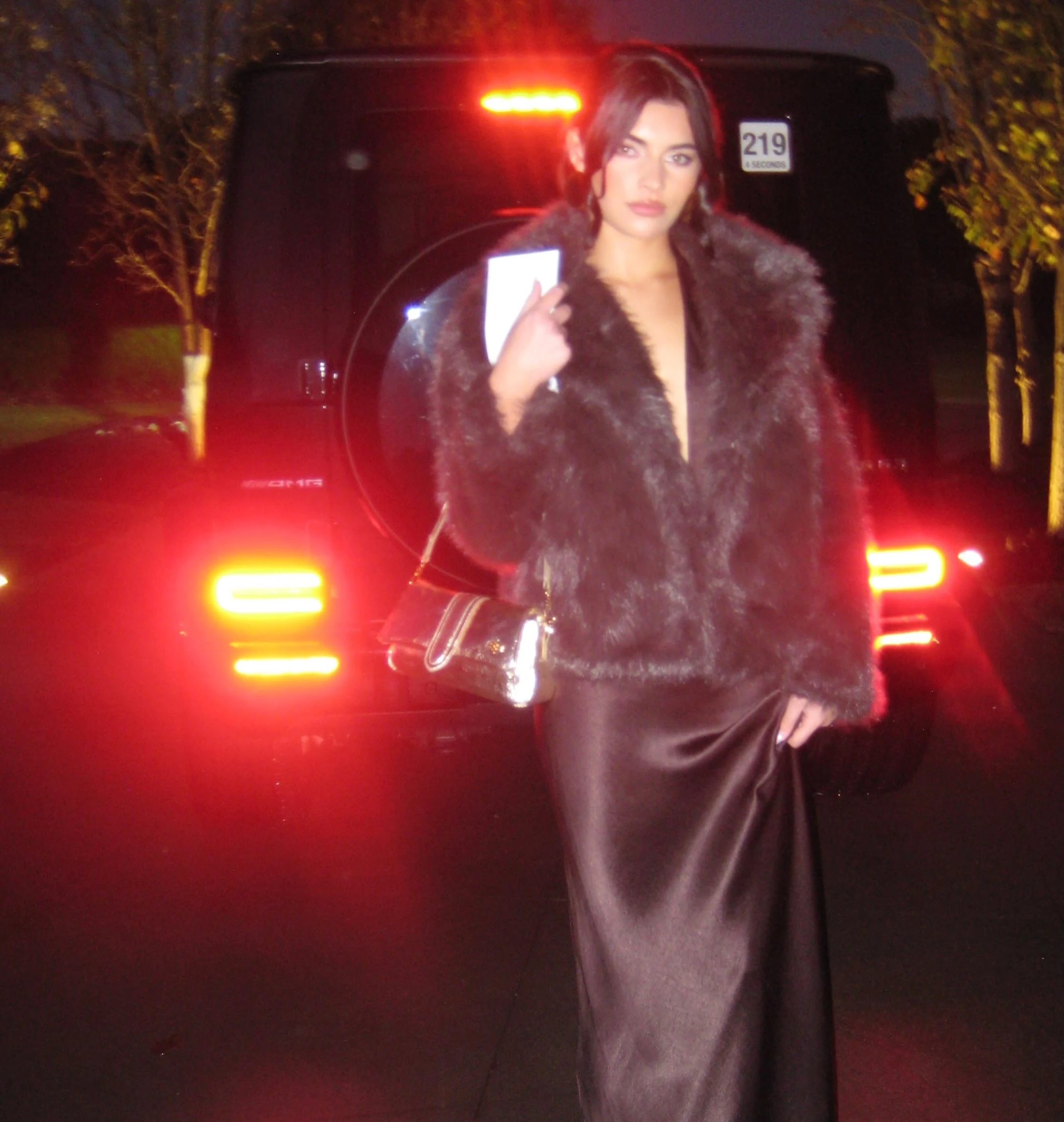Dark gray fur jacket layered over a long fitted dark brown slip dress with a small shoulder bag.