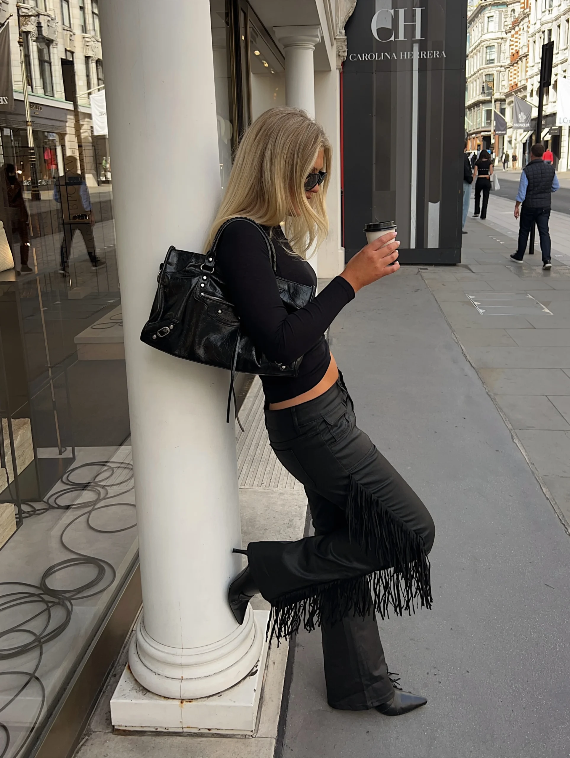 Fitted long-sleeve black crop top, high-waisted black leather flared pants with long fringe down outer legs, pointed black boots, large black patent-leather handbag, sunglasses