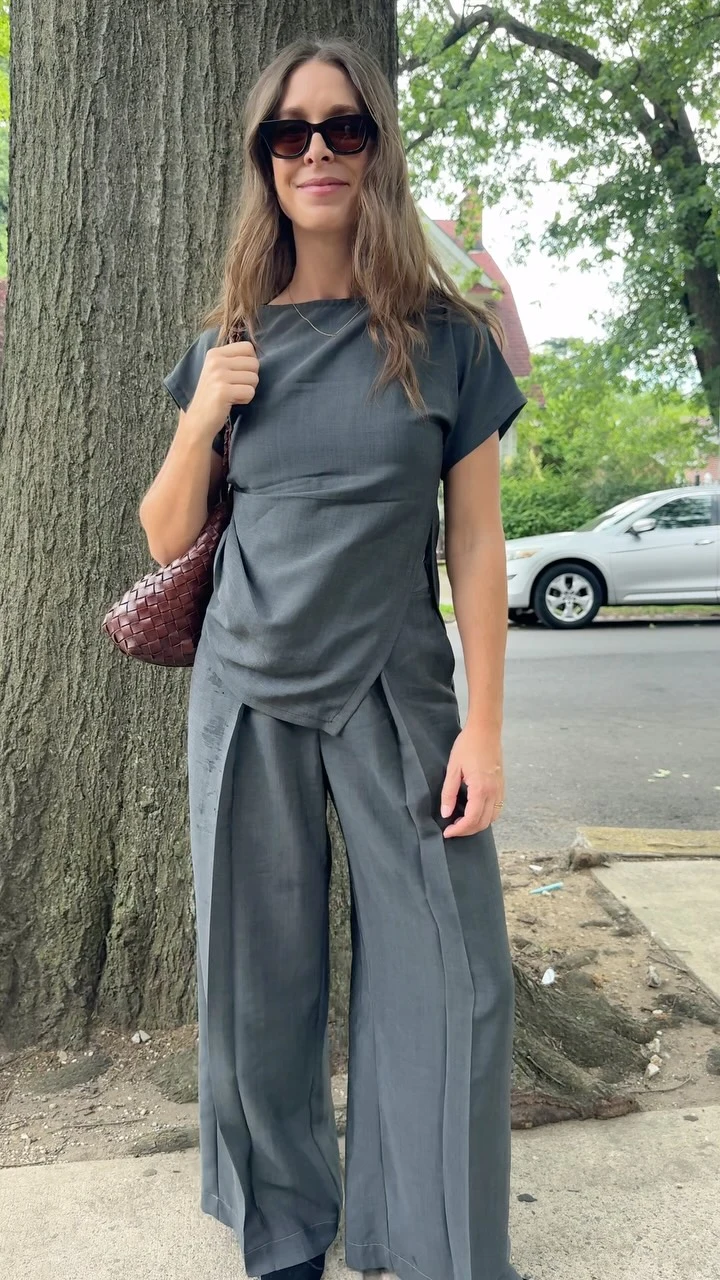 Person in matching charcoal-gray draped top and pleated trousers with brown woven bag and oversized sunglasses.
