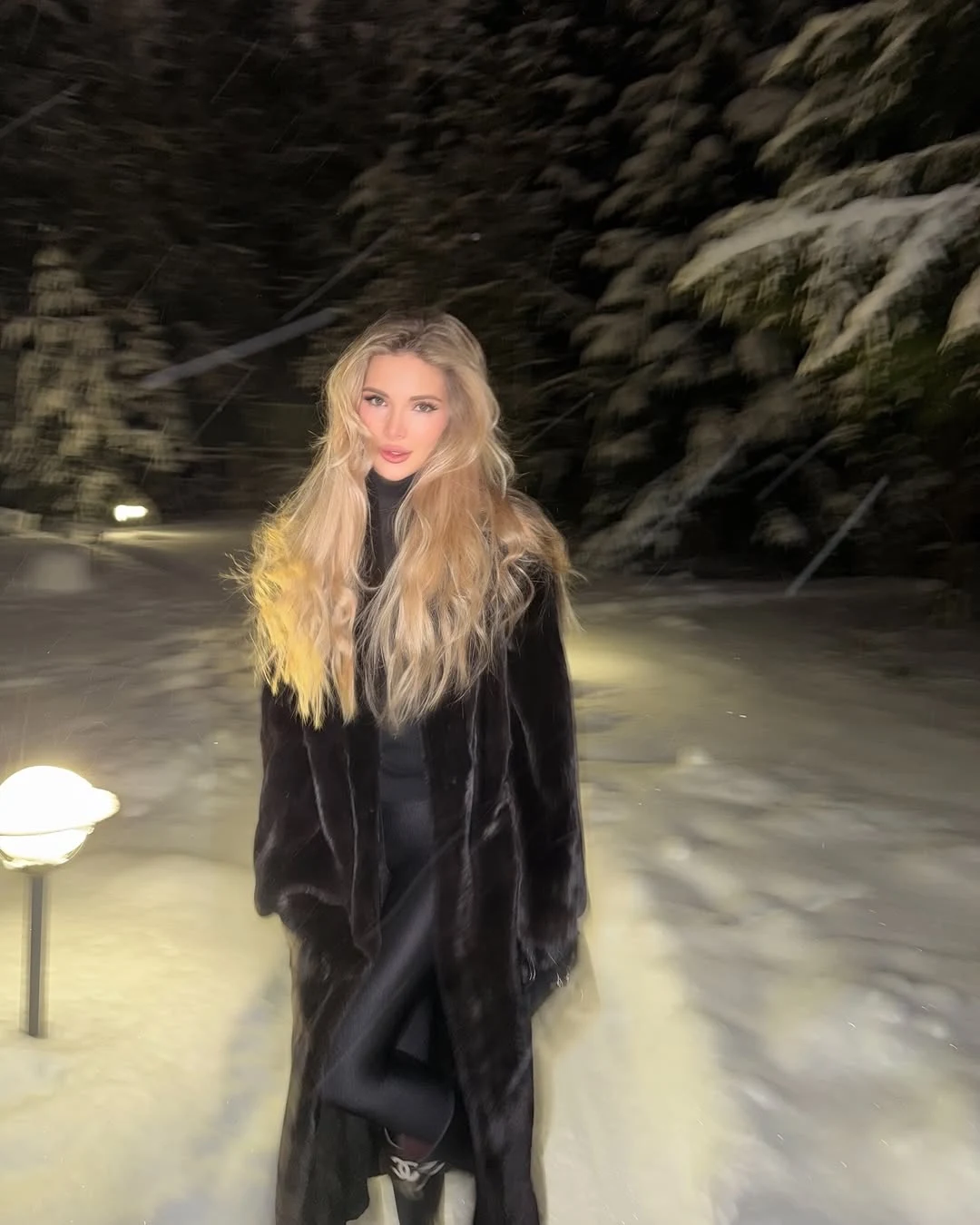 Woman in a long black fur coat, black tights, boots, headband, gloves, and a small black handbag.