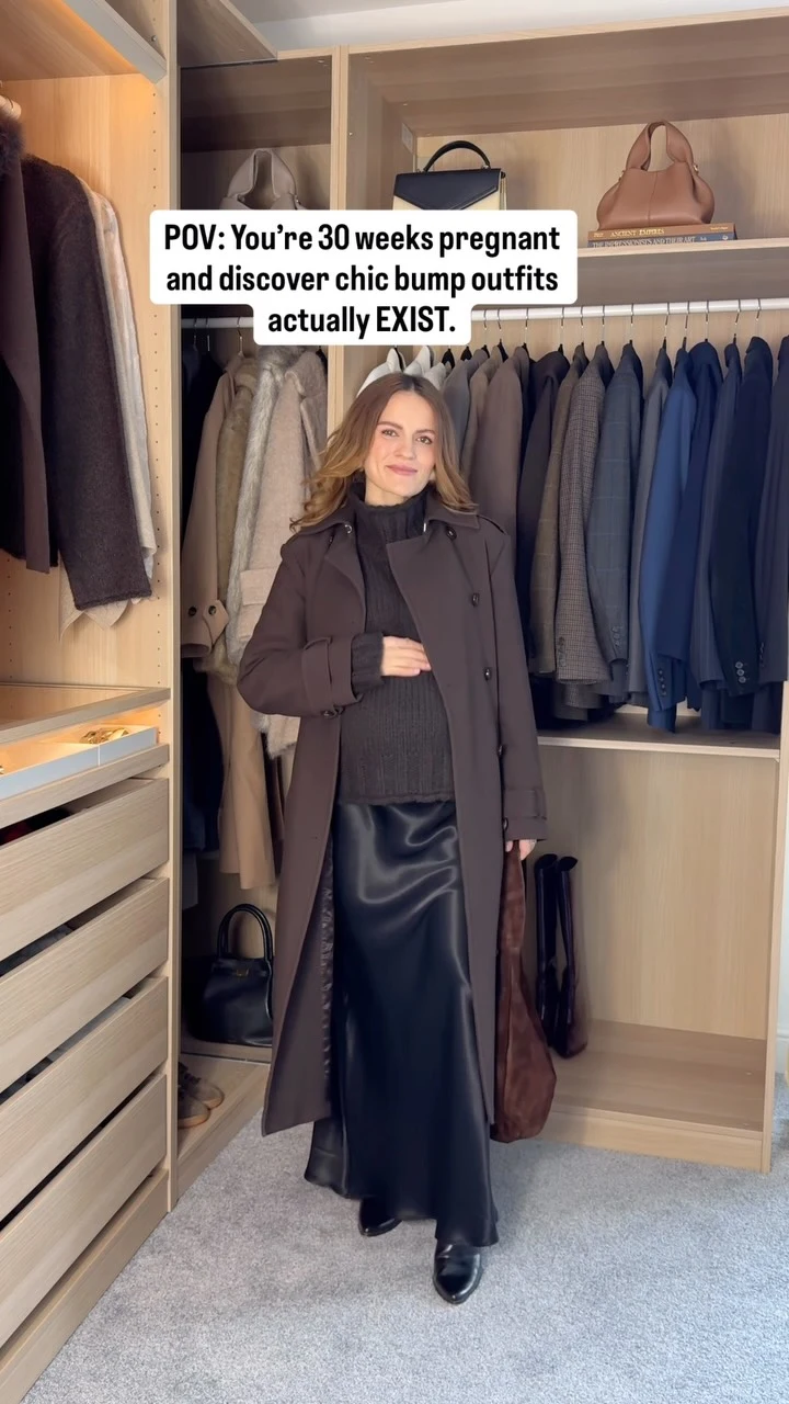 Pregnant woman in a long brown coat over a dark ribbed sweater and glossy black skirt in front of a closet.