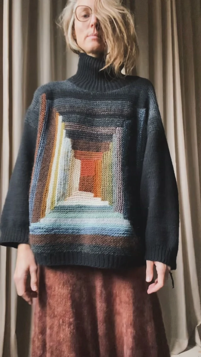 Person in a geometric-print black turtleneck sweater over a long brown skirt, wearing round eyeglasses.