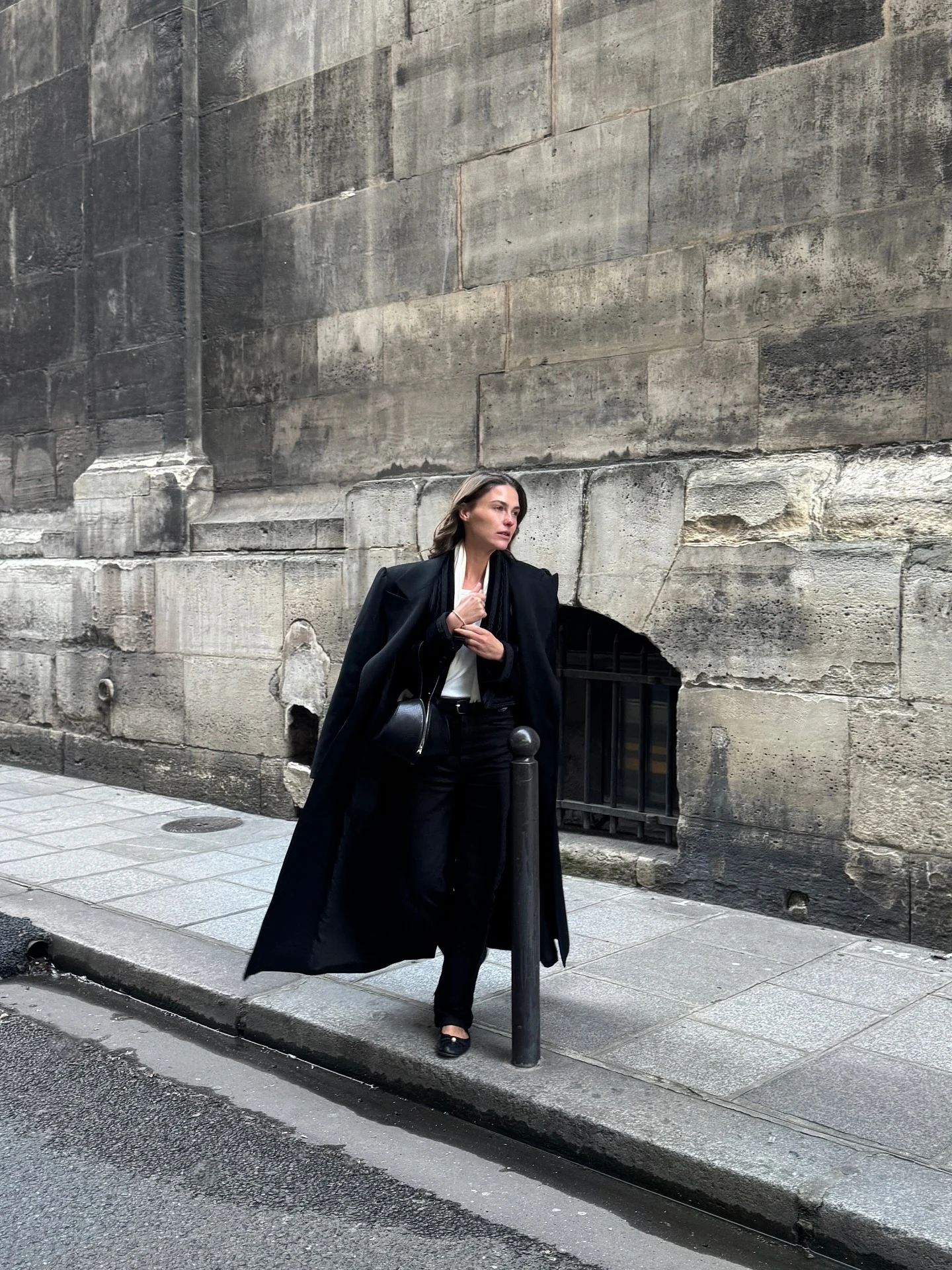 Woman in an oversized black wool coat, white top, straight trousers, quilted ballet flats, and small bag.