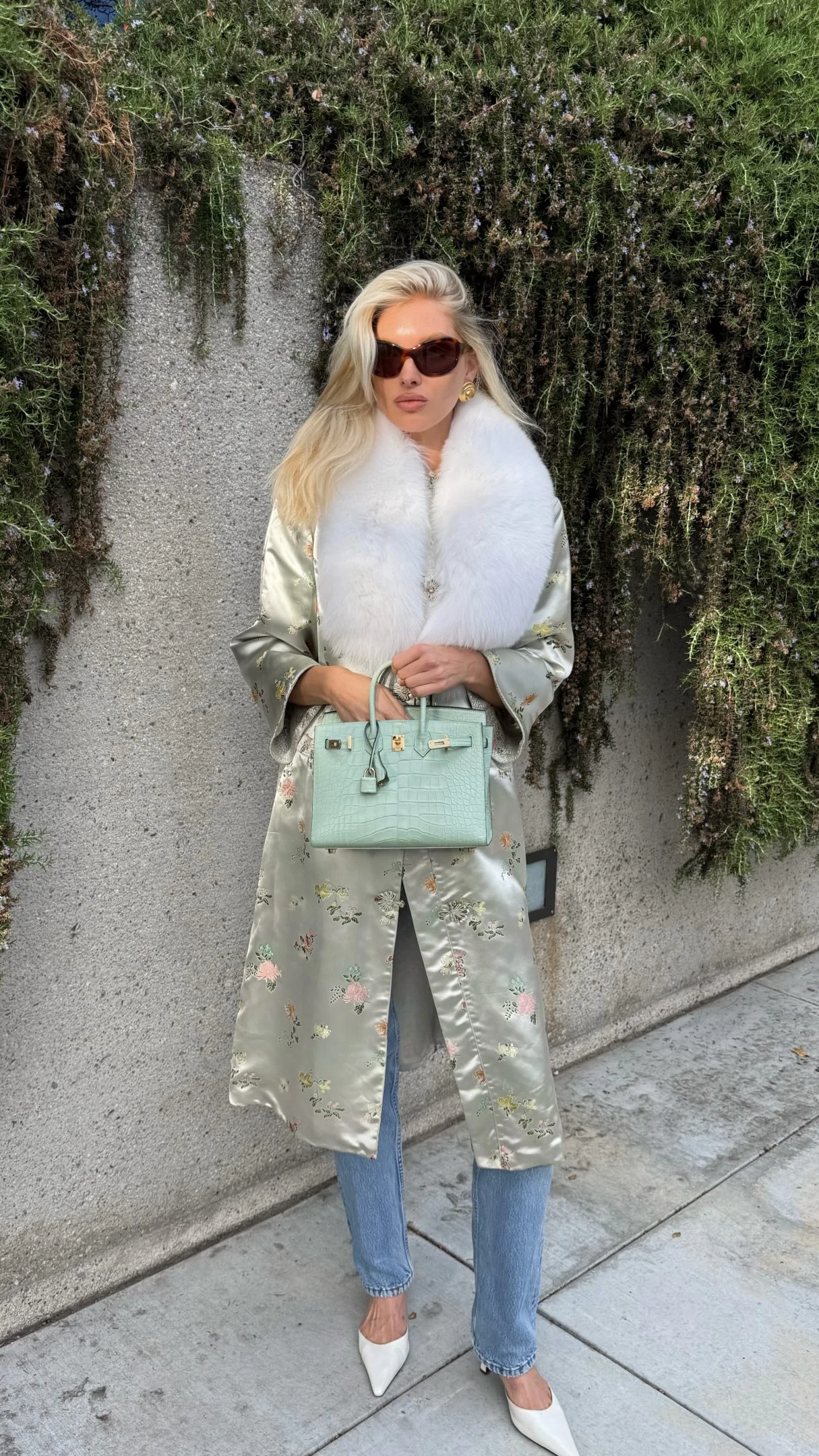Person in a pale-green floral satin coat, white faux-fur scarf, straight-leg jeans, and white slingback heels.