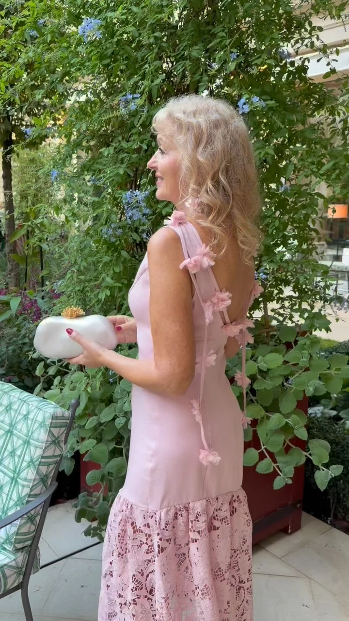 Pale pink sleeveless dress with a floral lace hem and small white clutch in a garden.