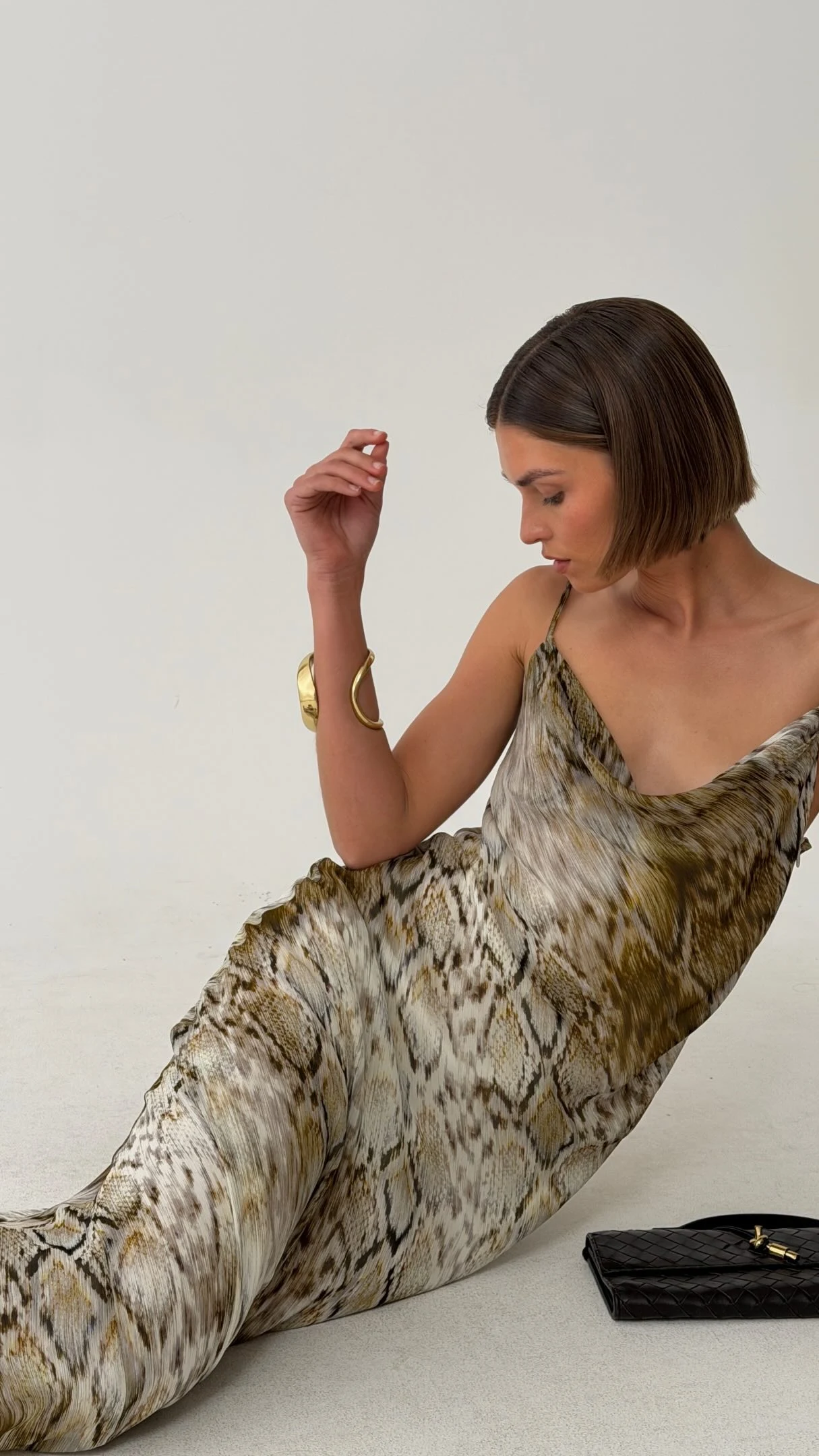 Long draped patterned dress with brown and olive tones, gold cuff, and black woven clutch.