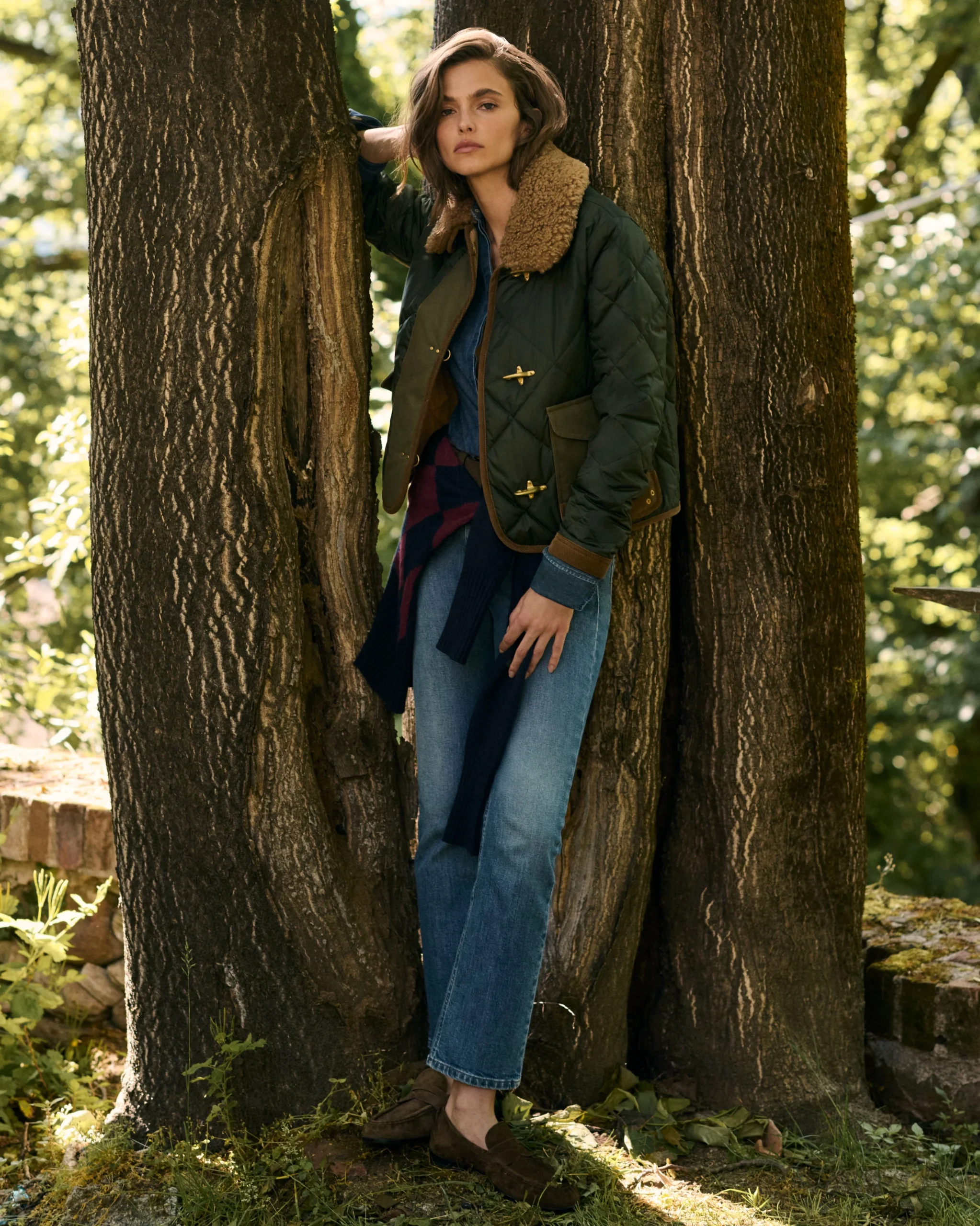 Person in dark green quilted jacket with tan collar and loose blue jeans standing between trees outdoors.