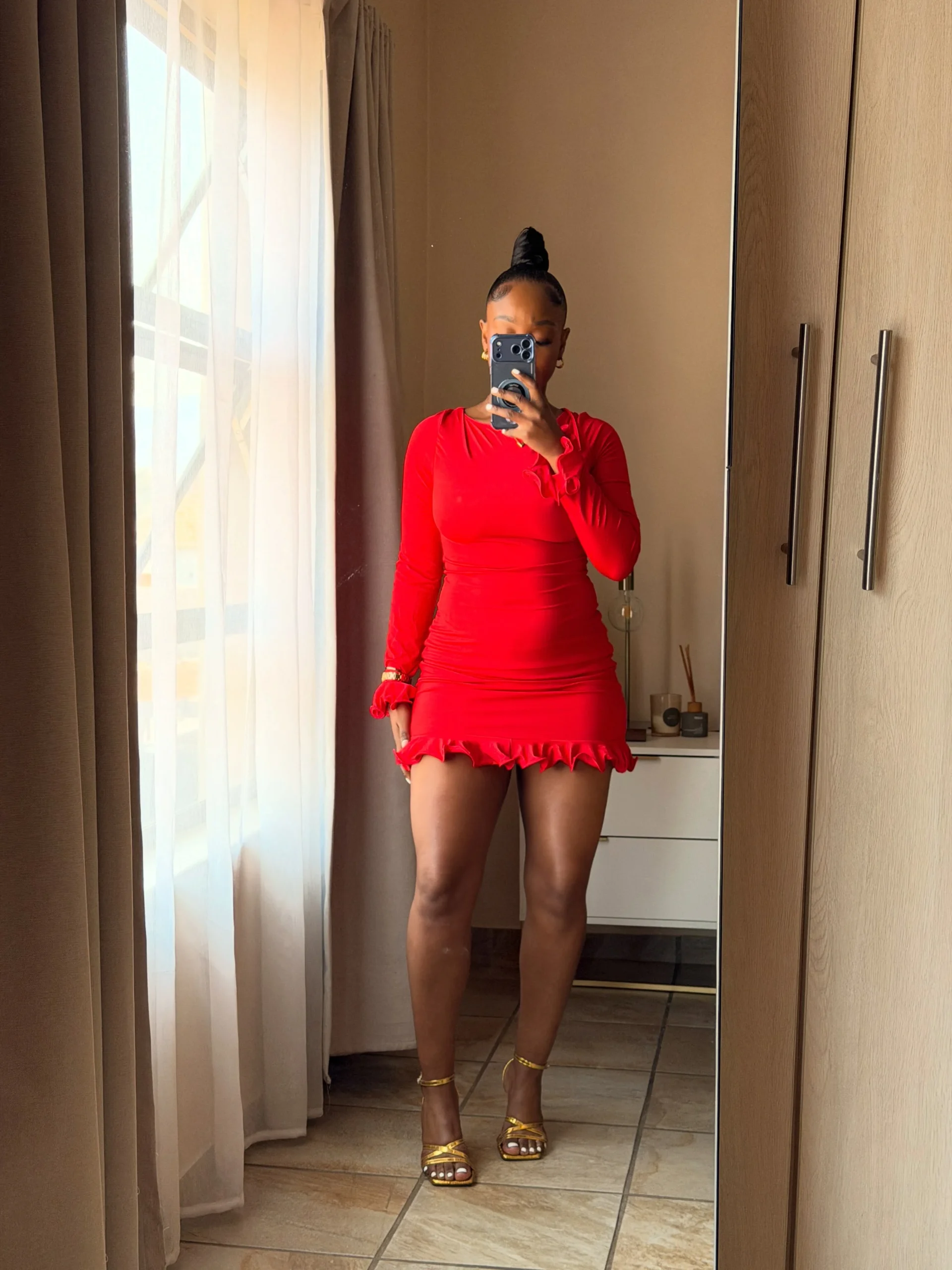 Woman in a vivid red ruched bodycon mini dress with ruffled cuffs and hem and gold strappy heels.
