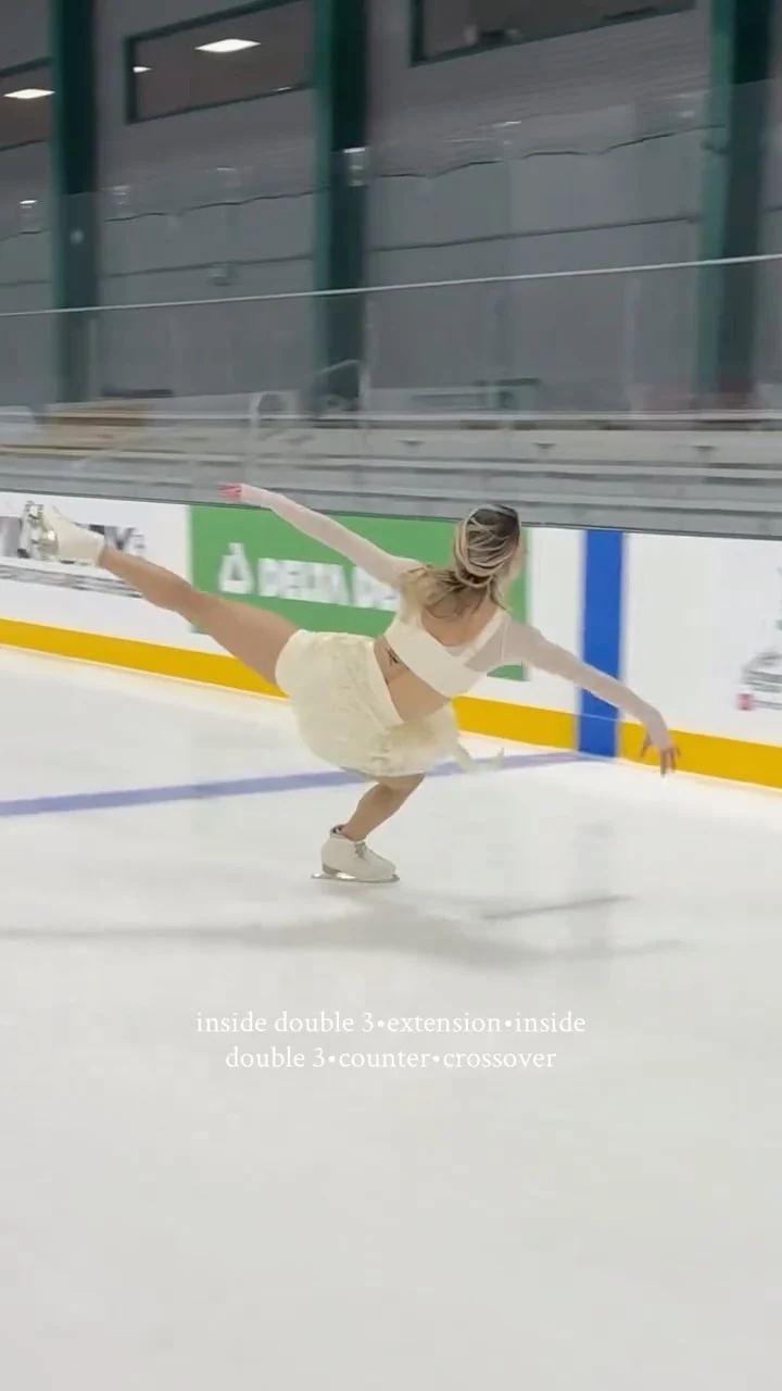Figure skater in a white long-sleeve top, flared skirt, and white skates gliding on an indoor rink.