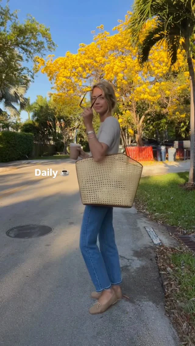 Person in white tucked top, blue straight-leg jeans, tan flats, and woven tote on a paved path.