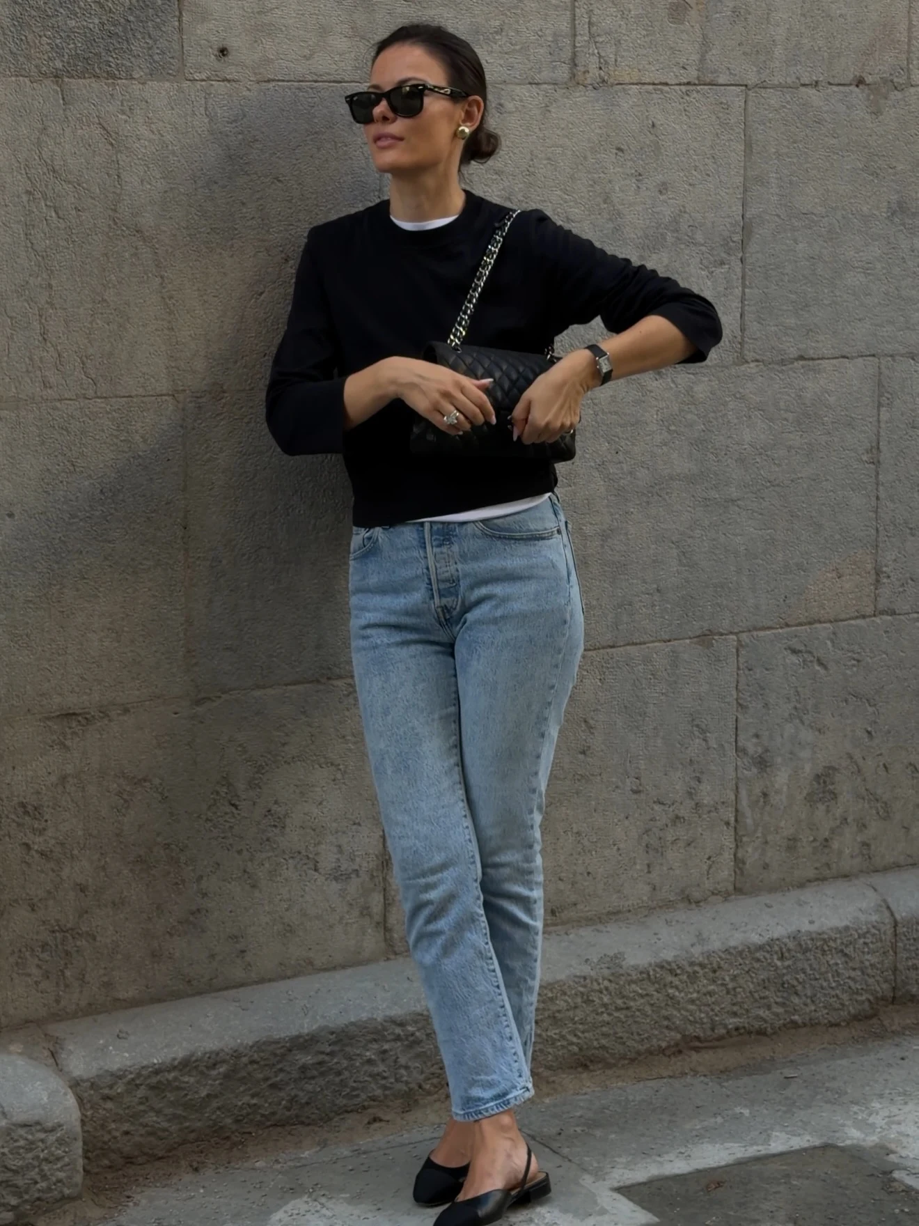 Black sweater, light blue straight-leg jeans, quilted bag, and black flats against a stone wall.