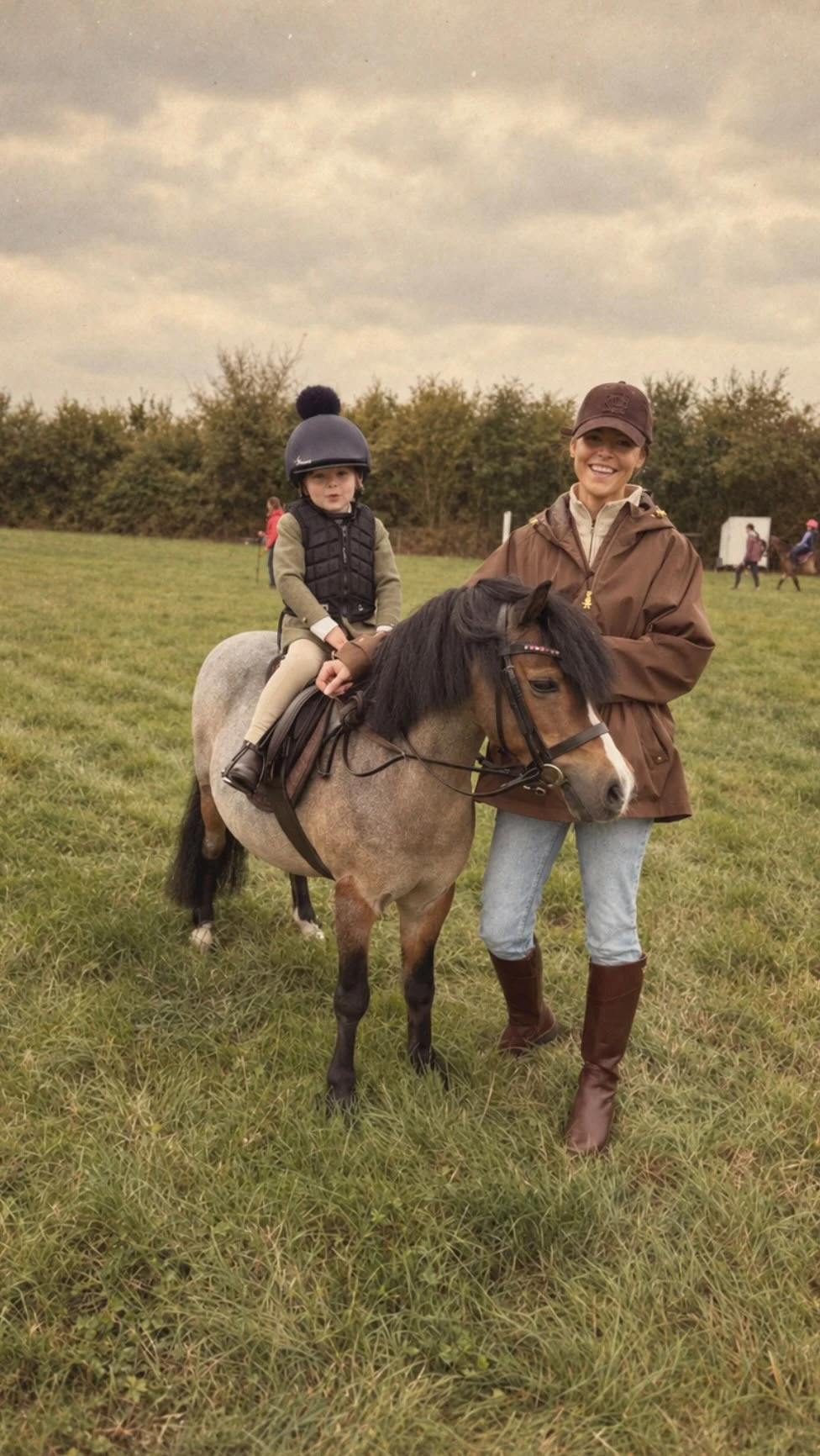 Adult in brown jacket and riding boots stands beside a child on a gray pony outdoors.