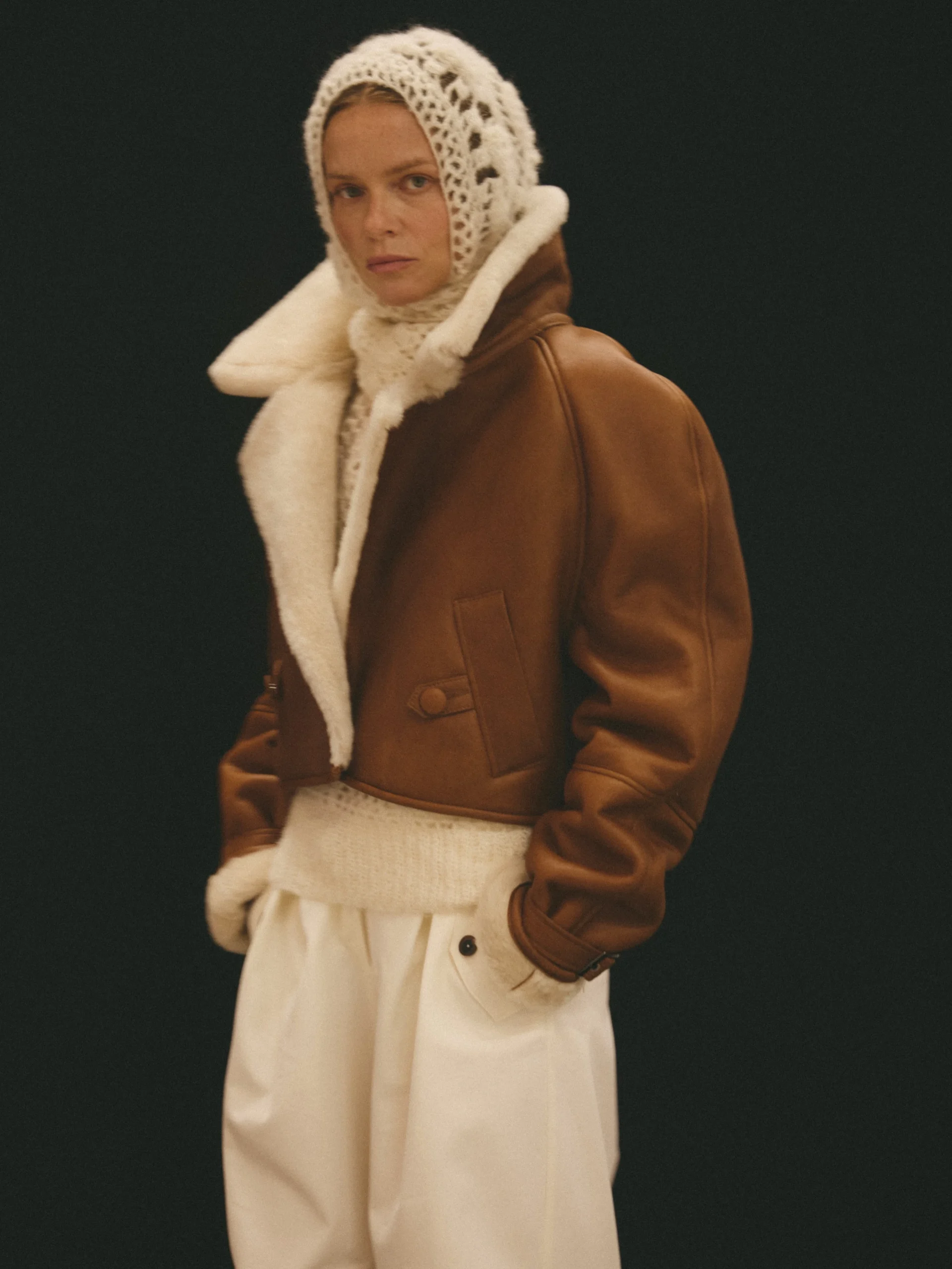 Cropped brown shearling jacket over cream knit top, loose cream trousers, and a cream openwork hood.