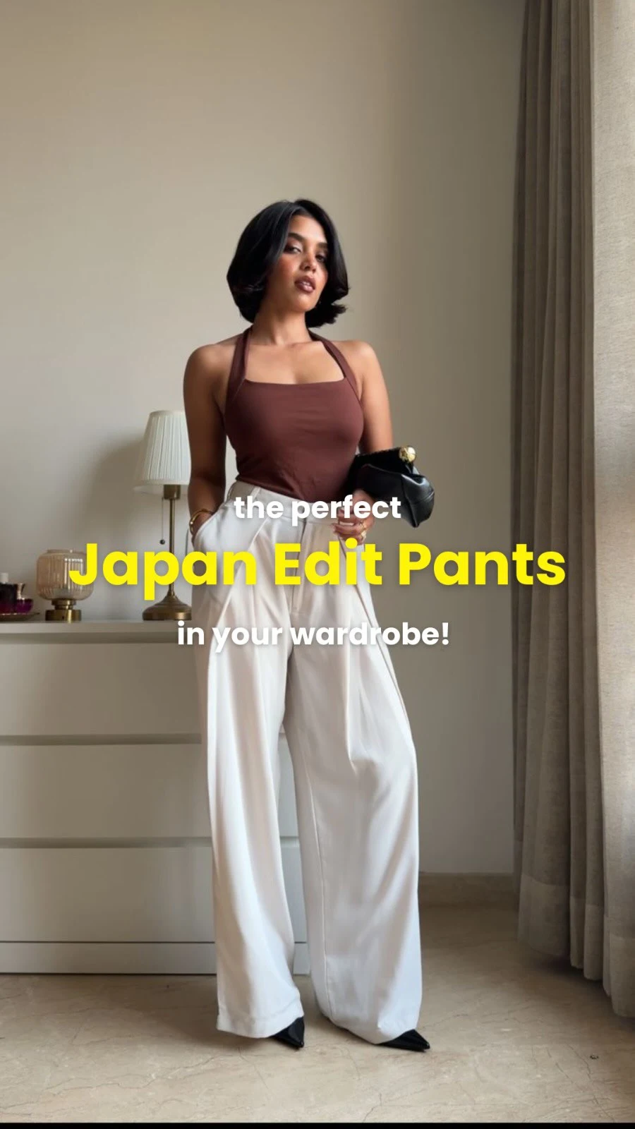 Brown sleeveless top tucked into white pleated trousers with black pointed shoes and a dark handbag.
