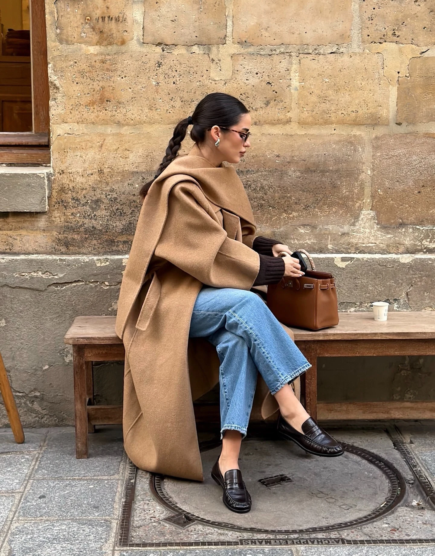 Seated person in a long camel coat, light blue jeans, black loafers, and dark sunglasses.