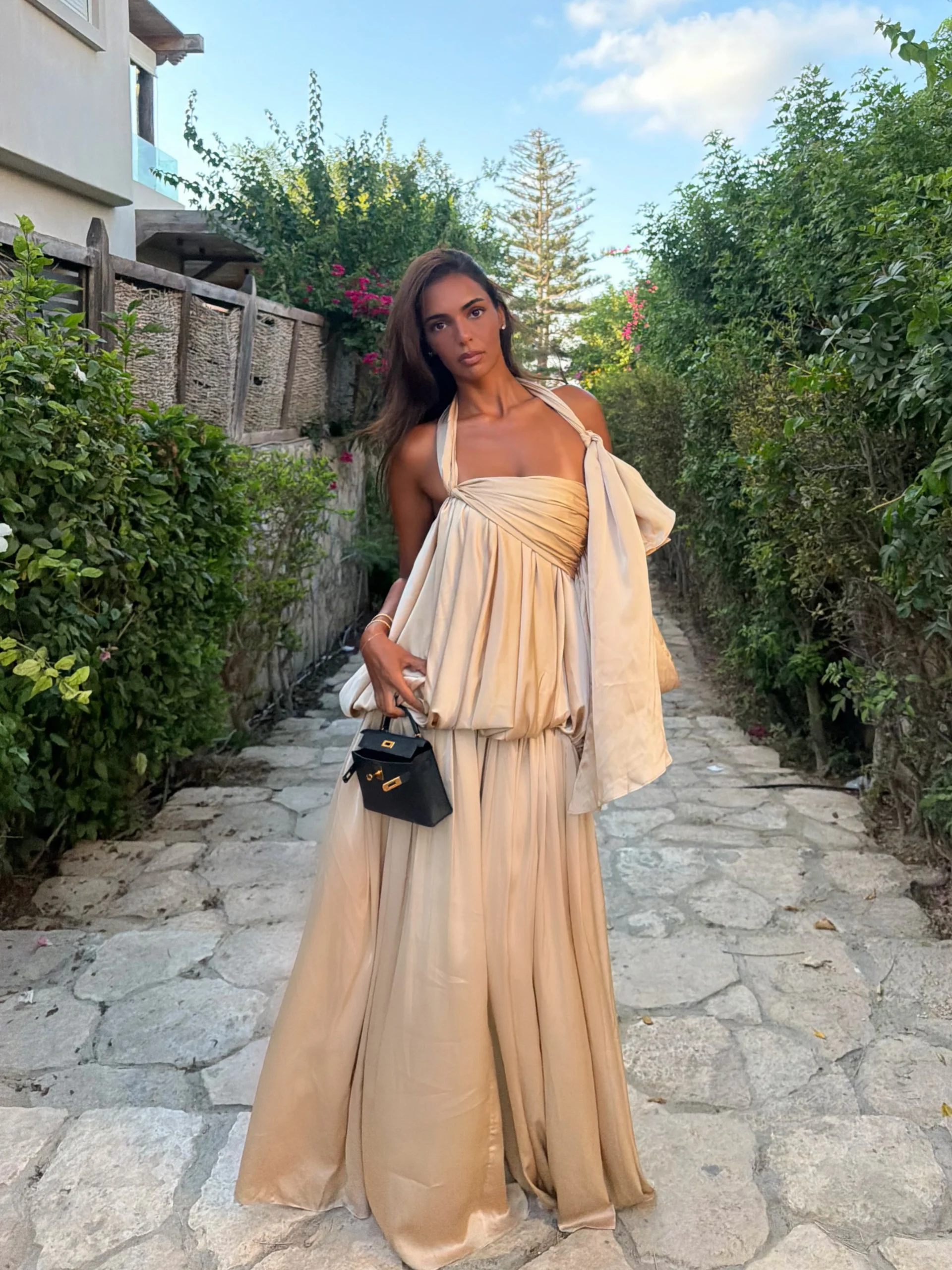 Floor-length champagne halter gown with draped bodice, flowing skirt, and small black structured handbag.
