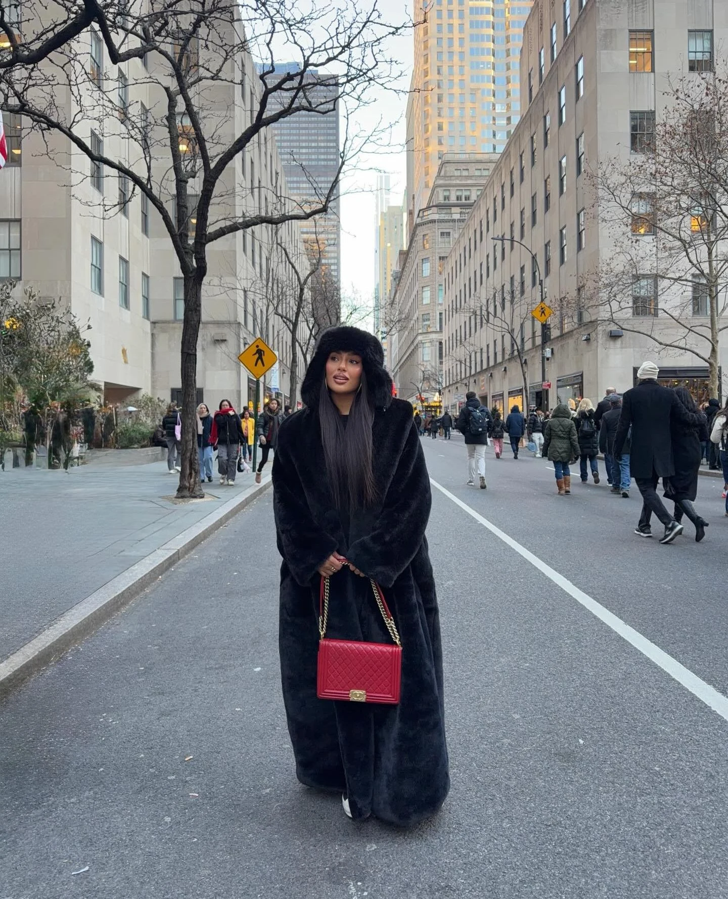 Woman wearing a floor-length black hooded faux-fur coat, red quilted bag, and black boots.