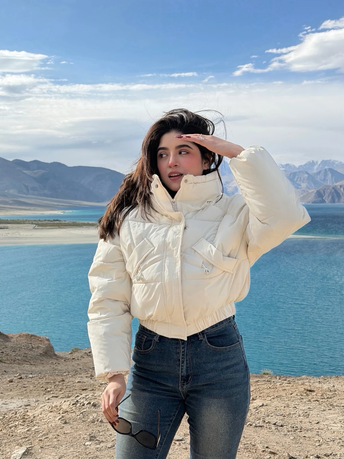 Woman in ivory cropped puffer jacket and dark blue high-waisted jeans holding sunglasses.