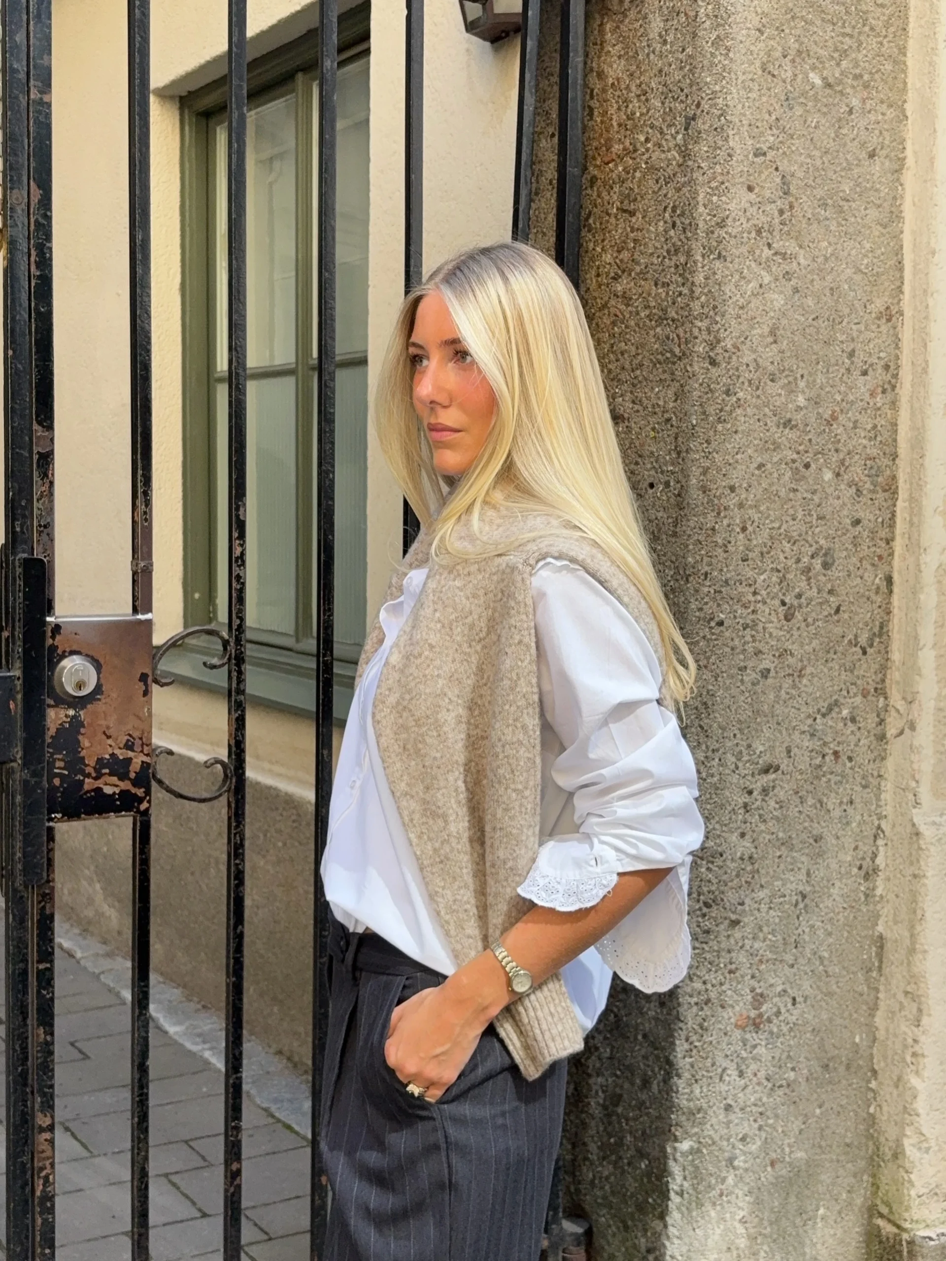 Light beige oversized knit sweater vest draped over shoulders, white long-sleeve button-up shirt with lace Peter Pan collar, dark gray pinstripe wide-leg trousers, black flip-flops