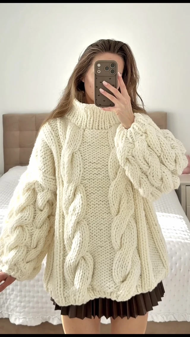 Person wearing a cream cable knit turtleneck over a dark brown pleated skirt indoors.