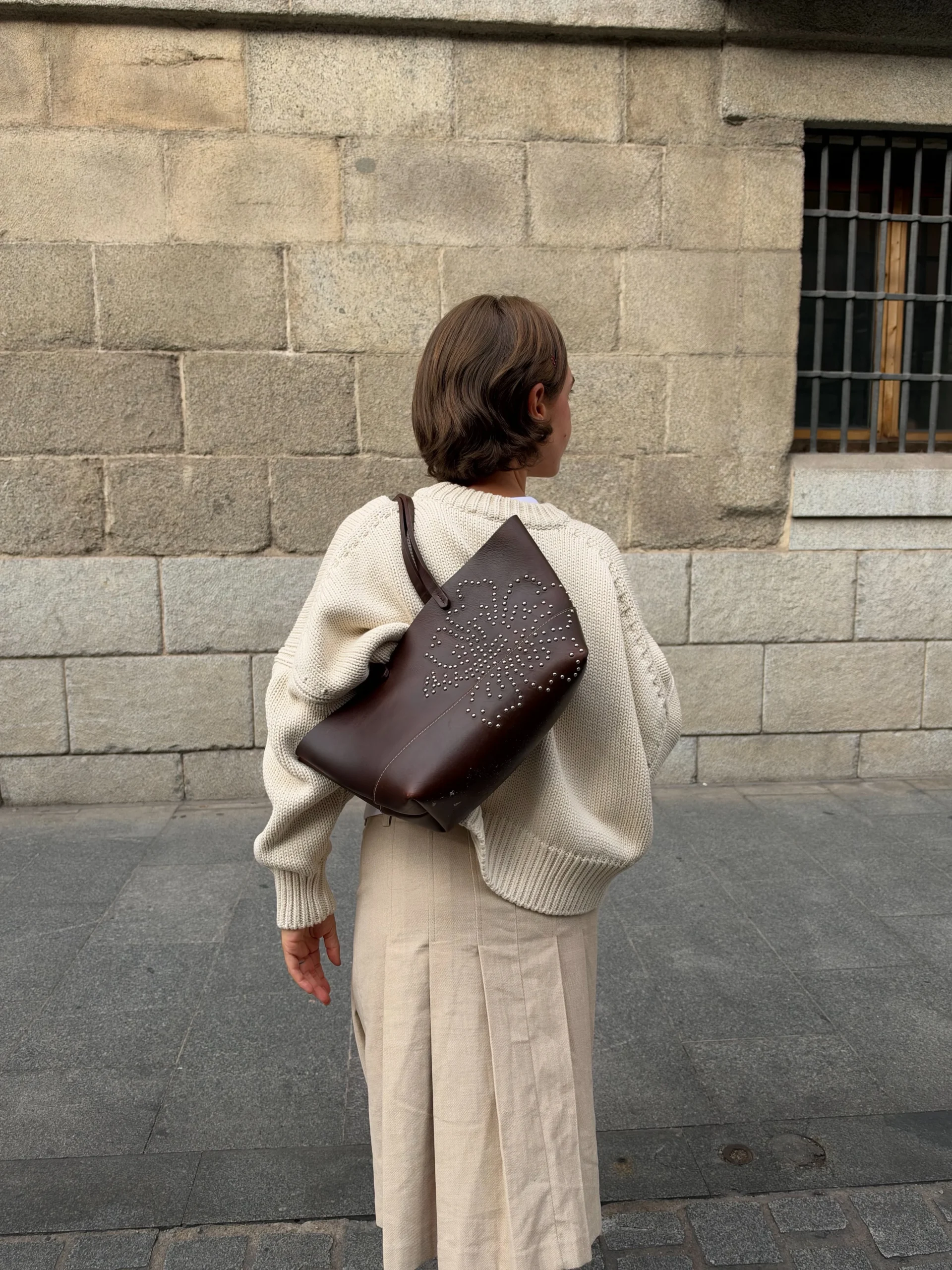 Oversized cream knit sweater, beige pleated midi skirt, white socks, brown clogs, and studded leather tote.